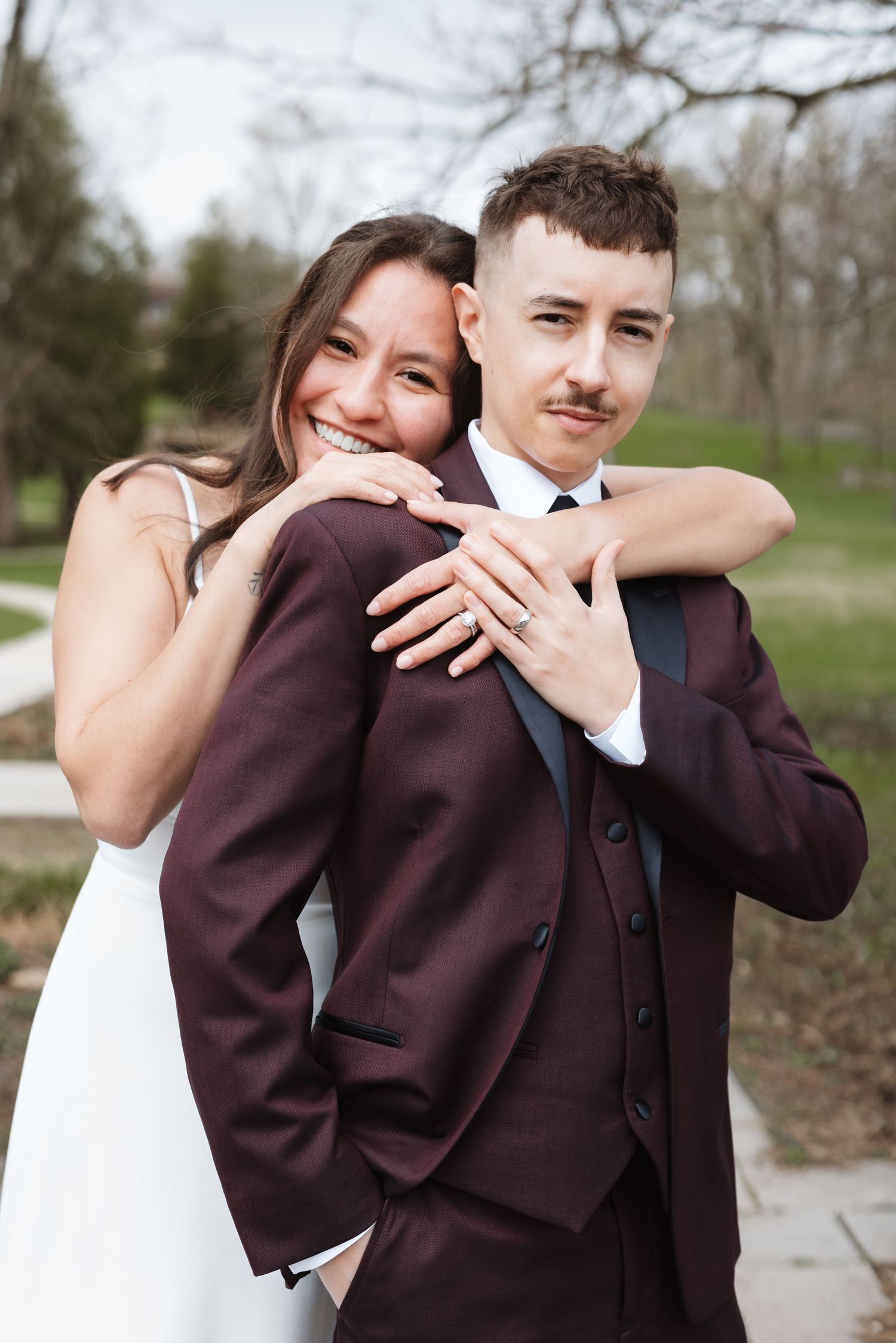 Bride in white dress hugs groom in burgundy suit from behind, both smiling outdoors at their wedding.
