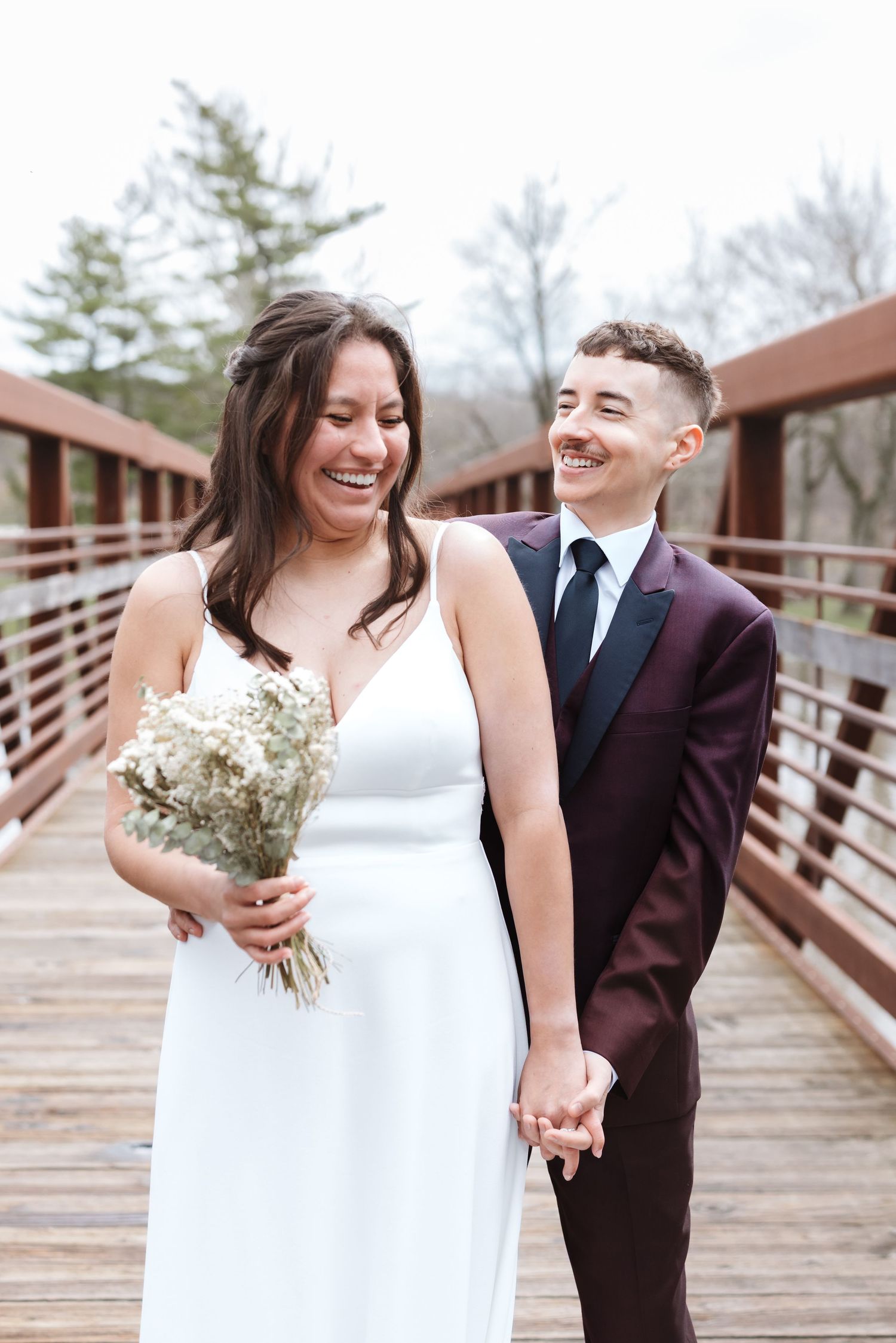 A laughing bride in a white dress holds a bouquet while a partner in a burgundy suit holds her hand on a wooden bridge.
