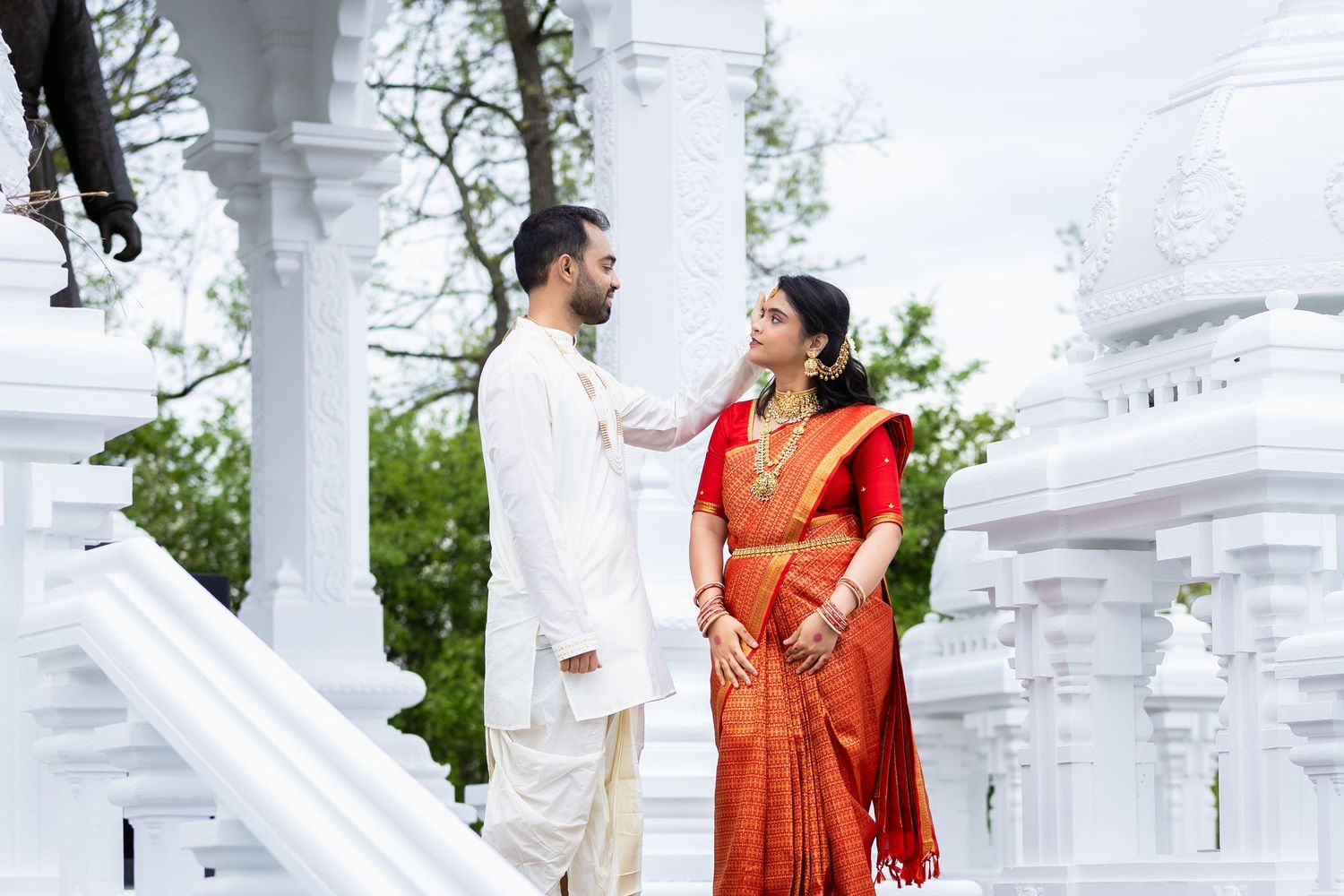 Indian bride in red silk saree and groom in white kurta posing at ornate white Hindu temple entrance.