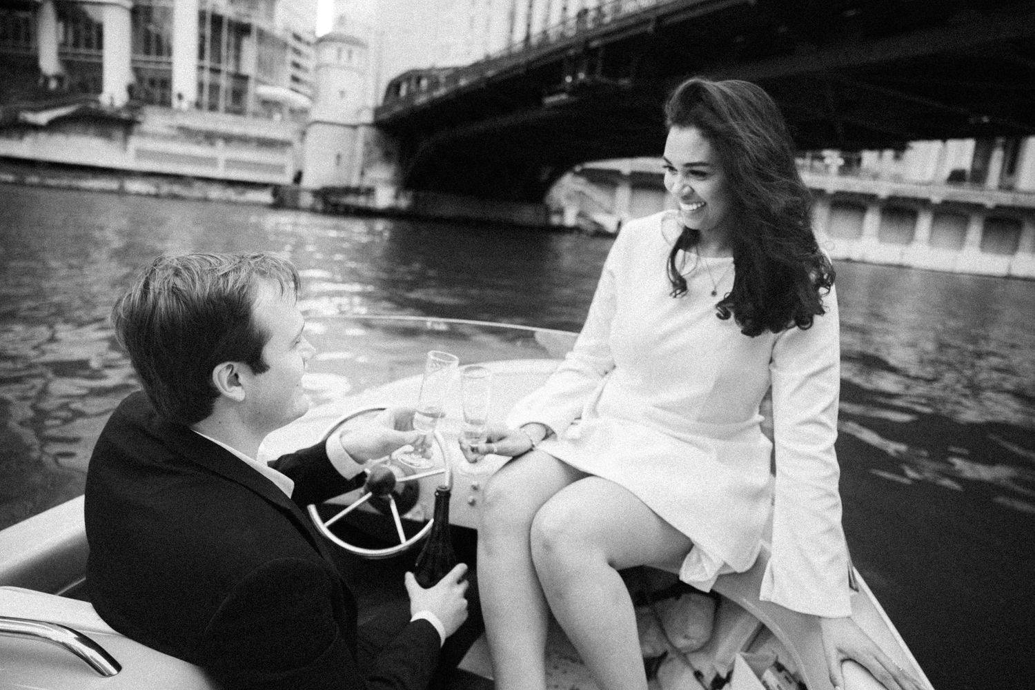 Black and white photo of engaged couple laughing on a vintage boat along Chicago river with bridge backdrop.