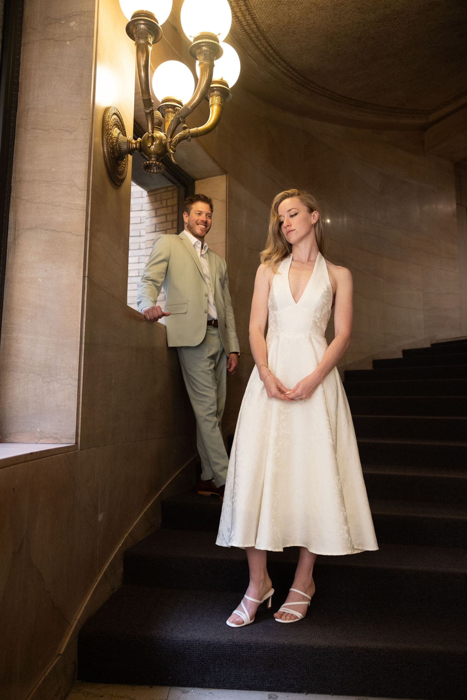 Bride in white midi halter dress poses on stairs as groom in sage suit smiles behind her.