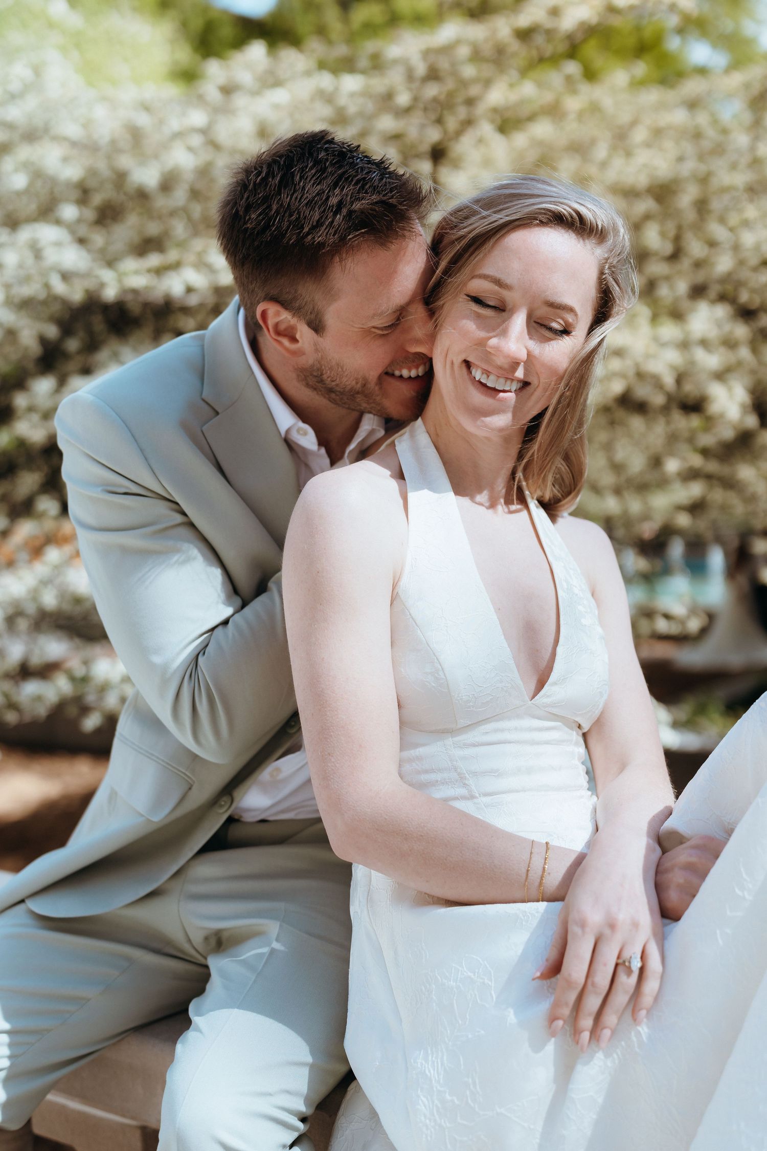 Groom whispers to laughing bride while seated outdoors among white blossoming trees.