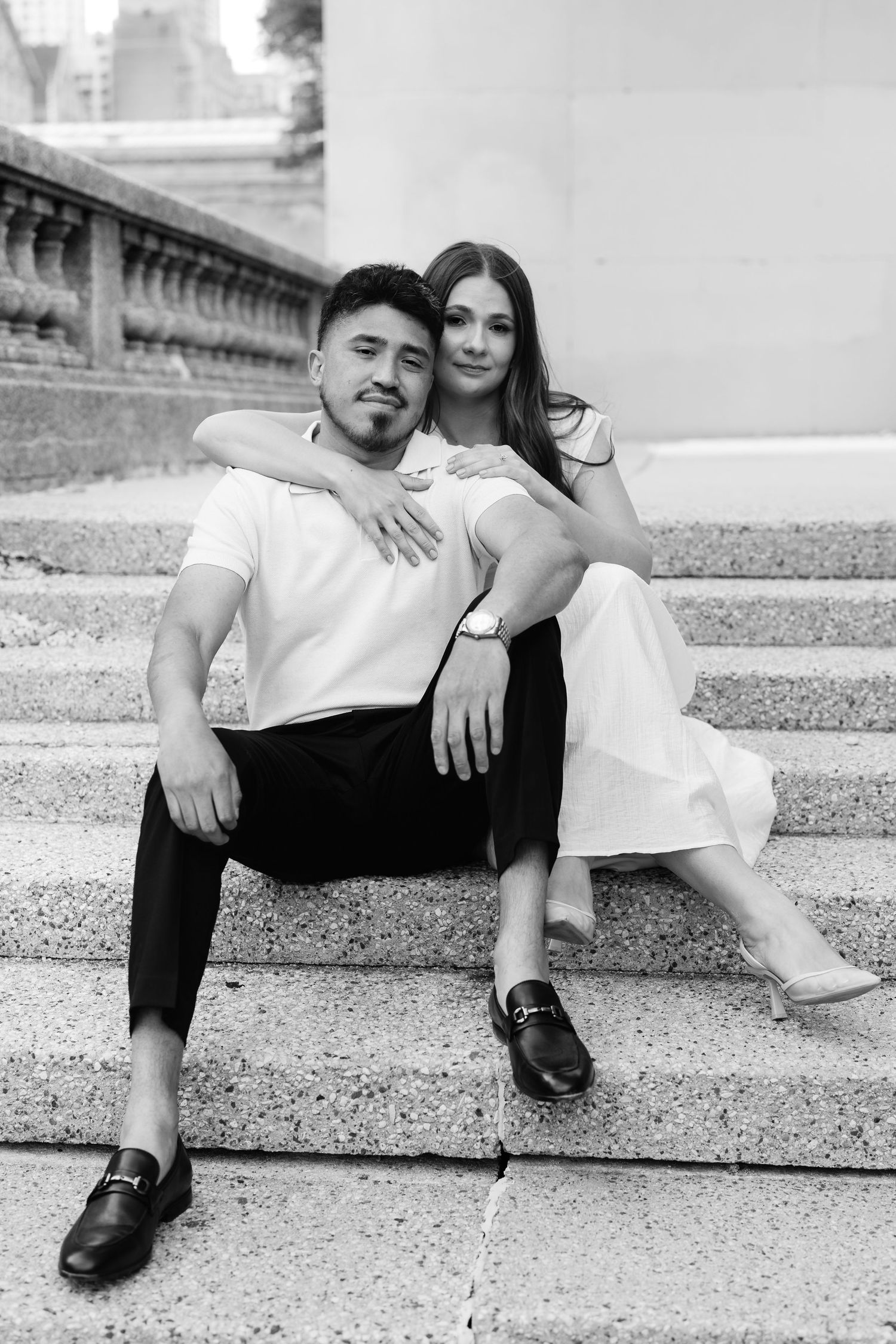 Black and white photo of couple sitting on stone steps, woman hugging man from behind, both smiling at camera.