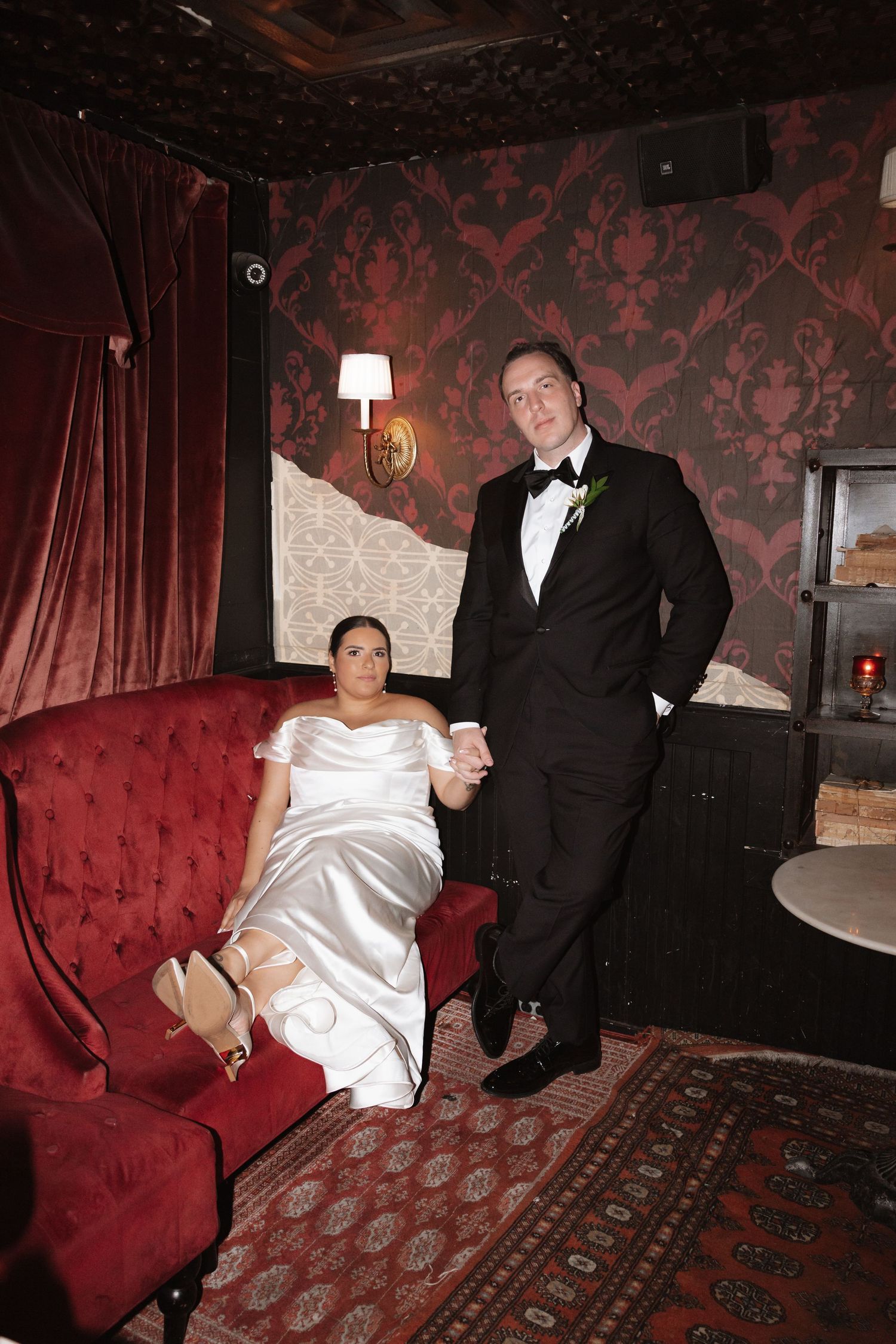 Bride and groom pose in elegant red velvet lounge, bride seated on tufted sofa holding hands.