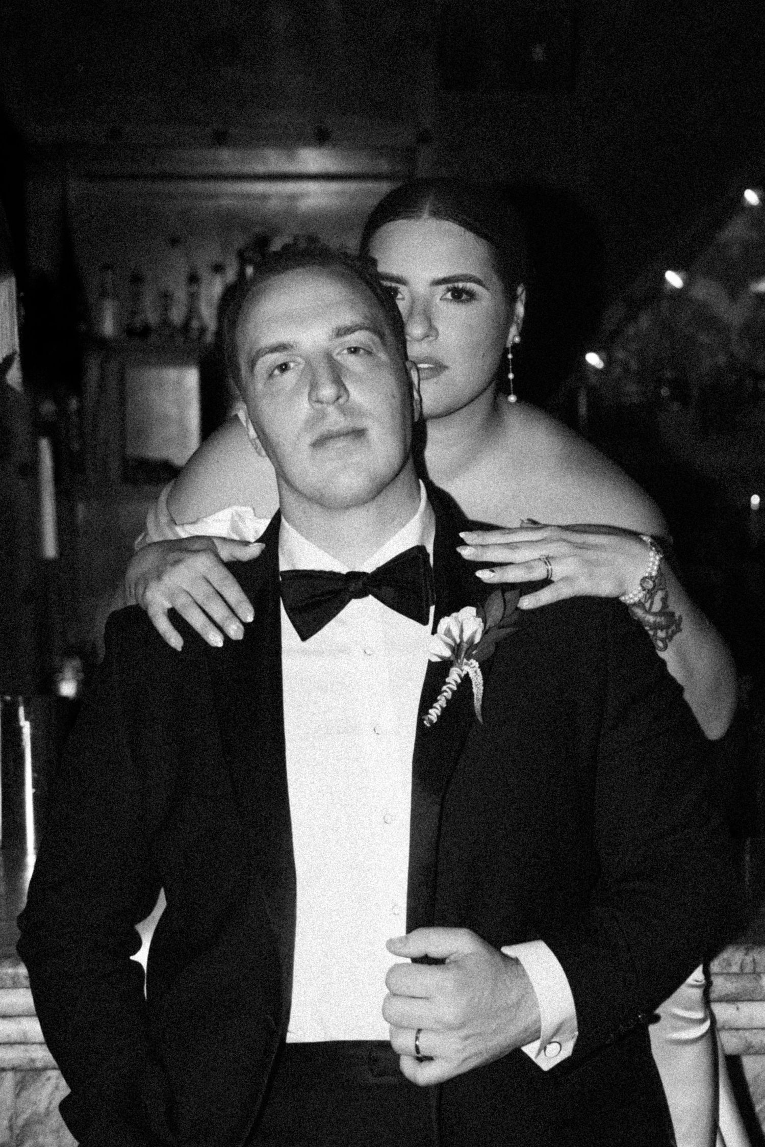 Black and white portrait of groom in tuxedo with bride embracing him from behind at wedding reception.
