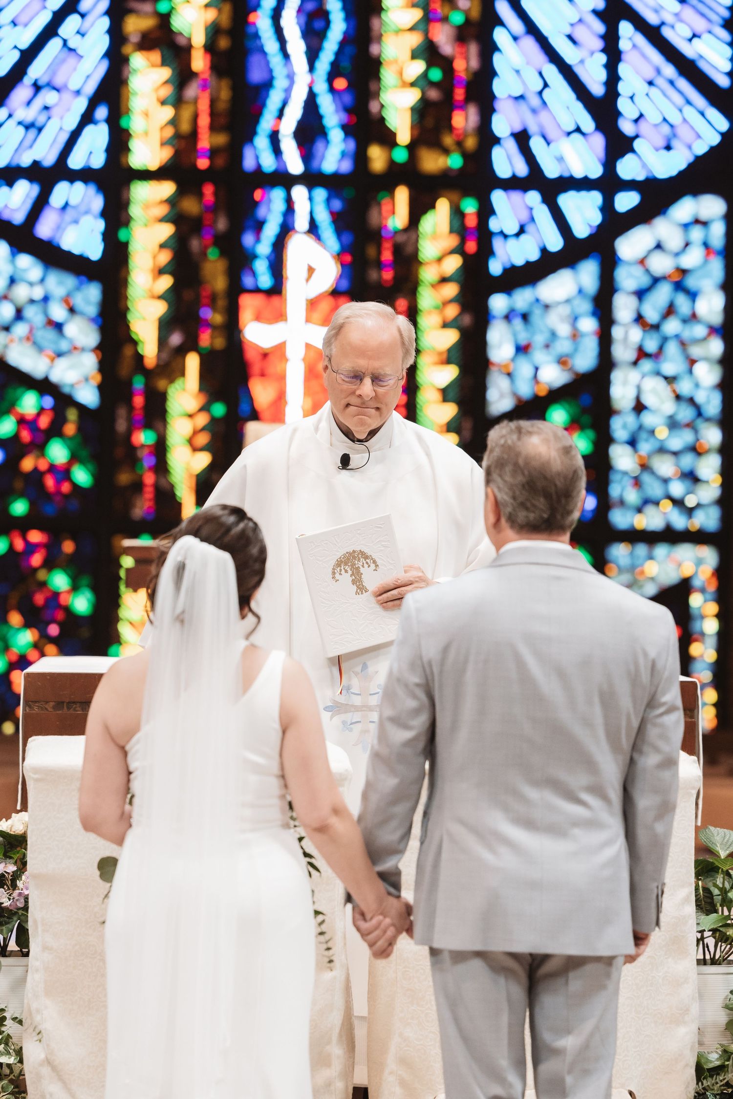 Bride and groom hold hands at church altar during wedding ceremony with colorful stained glass windows behind officiant.