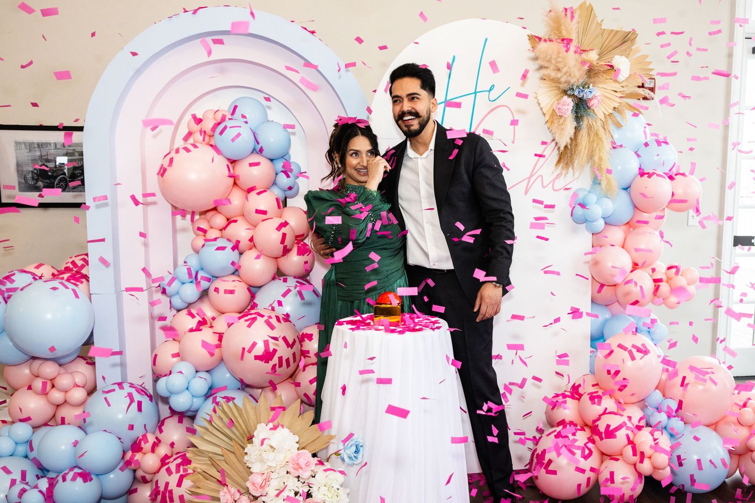 Smiling couple posing at gender reveal party with pink and blue balloon arch decor.