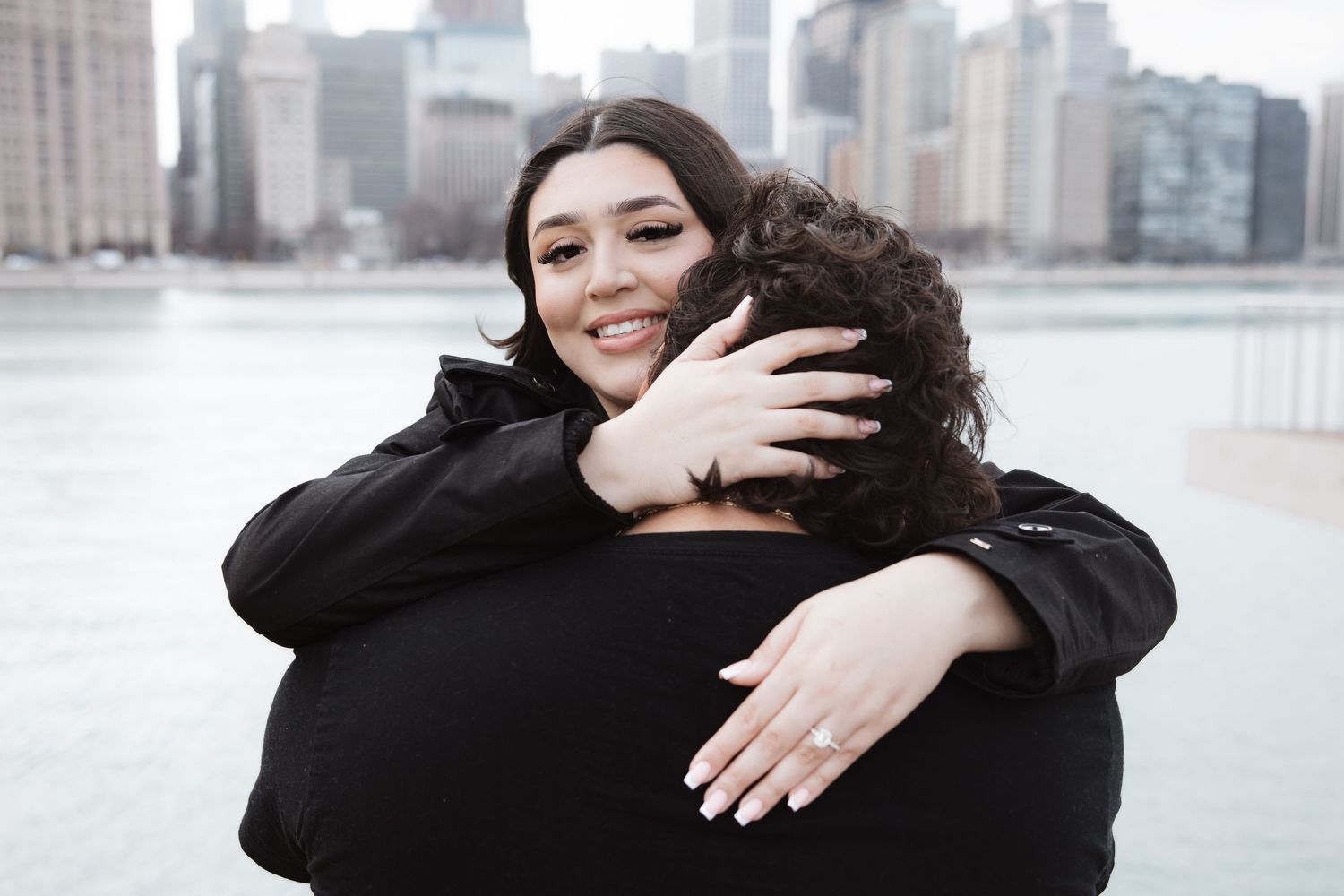 Joyful woman hugging her partner while showing off her engagement ring with the city skyline behind.