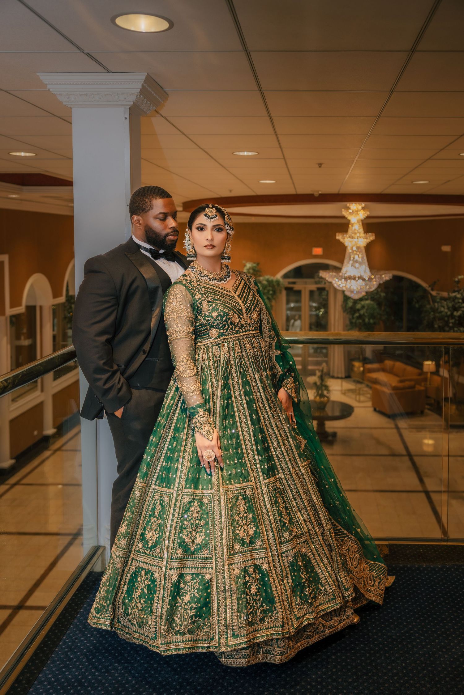 Bride in stunning emerald green and gold lehenga poses with groom in dark suit in elegant venue.