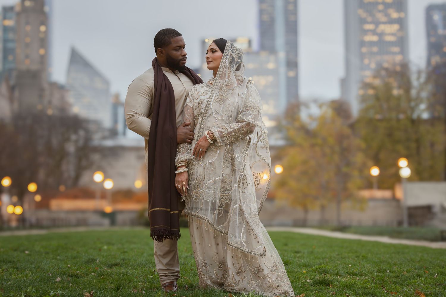 Interracial couple in traditional South Asian wedding attire pose in a park with city skyline backdrop at dusk.