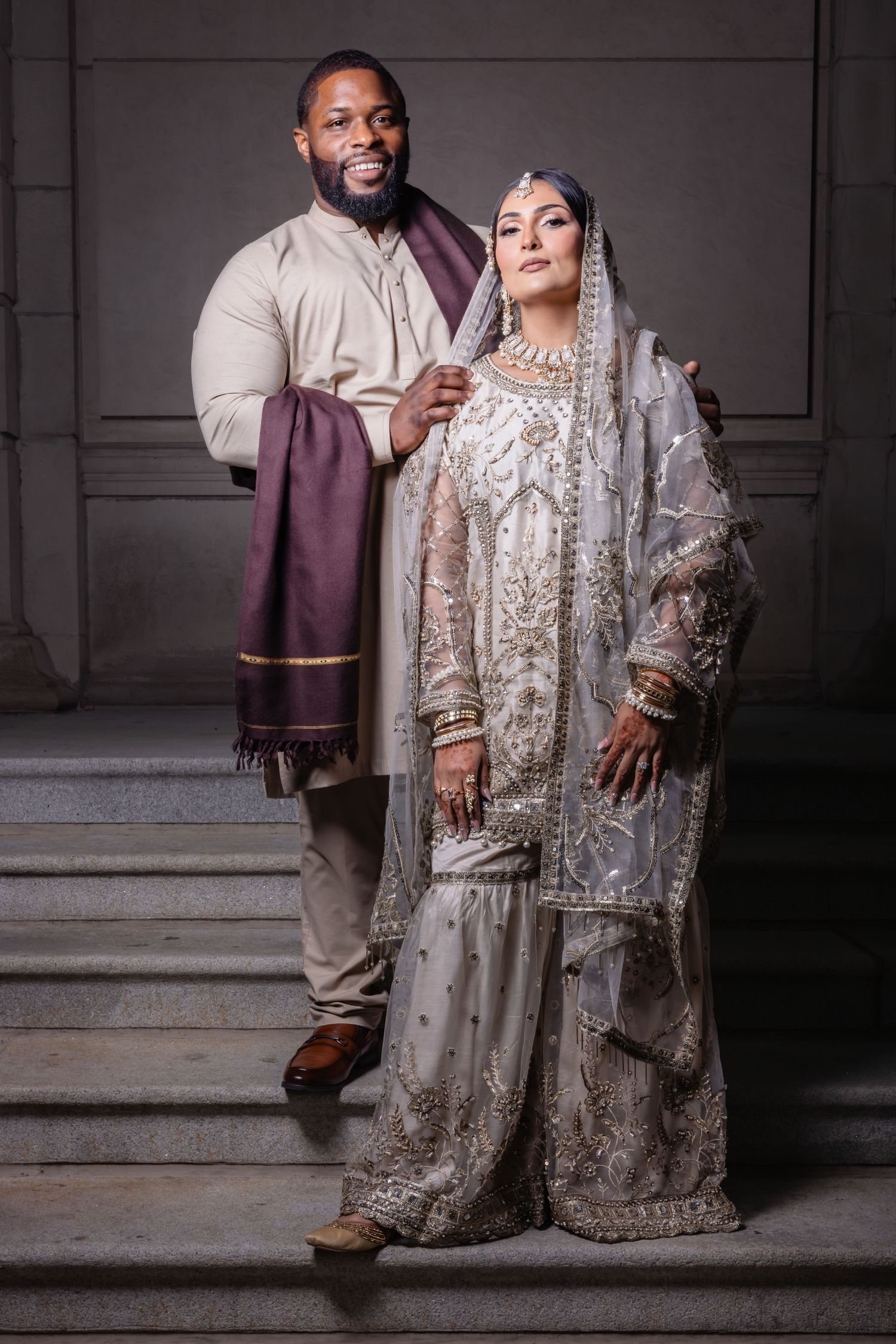 Groom in beige kurta holding purple shawl beside bride in silver embroidered lehenga on stairs.