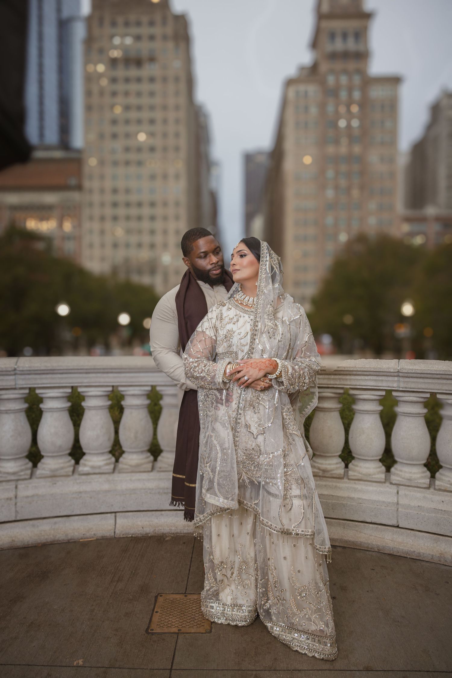 South Asian bride and groom in traditional wedding attire embrace on a stone balcony with city skyline.