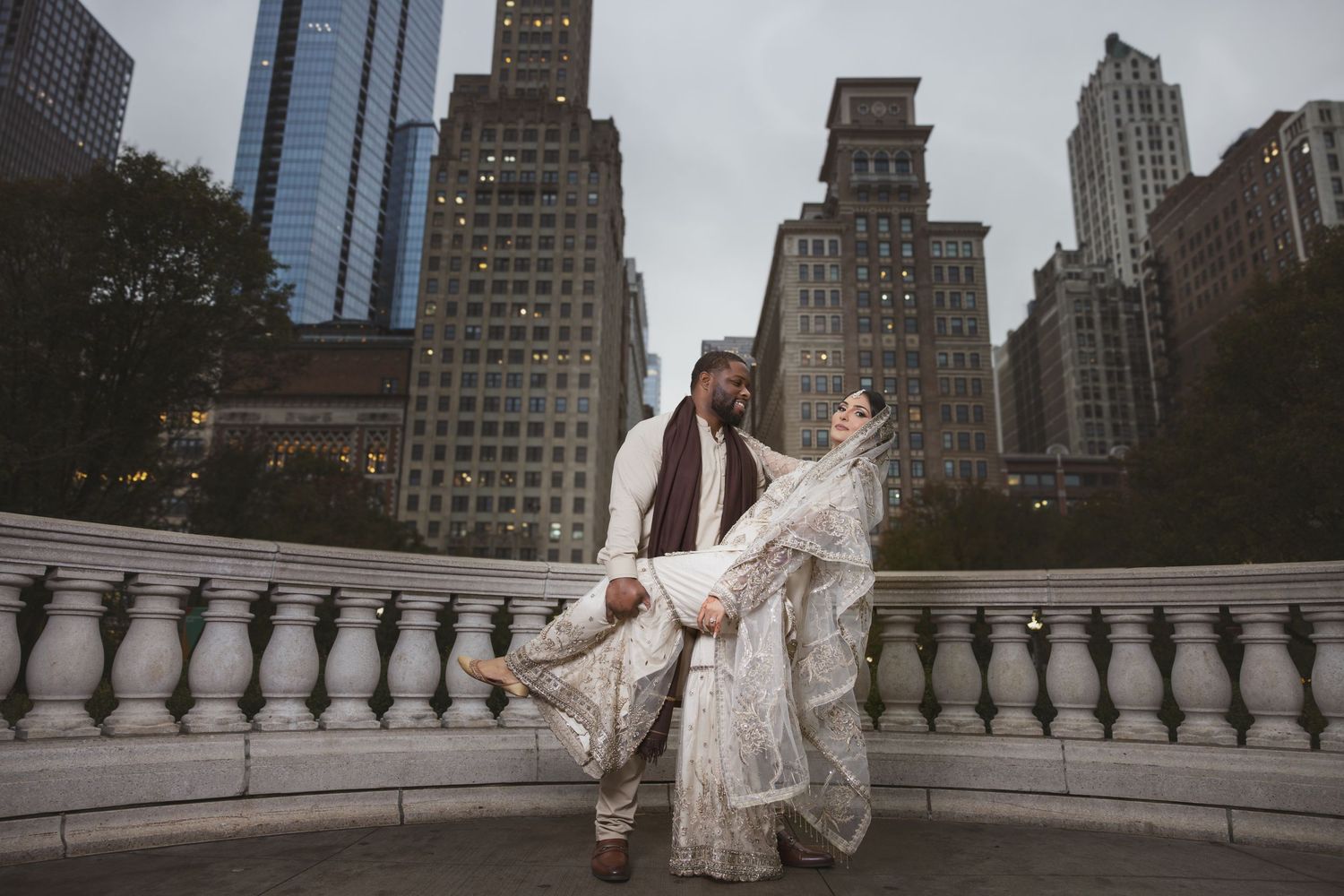 Groom lifting bride in ornate white lehenga at NYC landmark with city skyscrapers behind them.