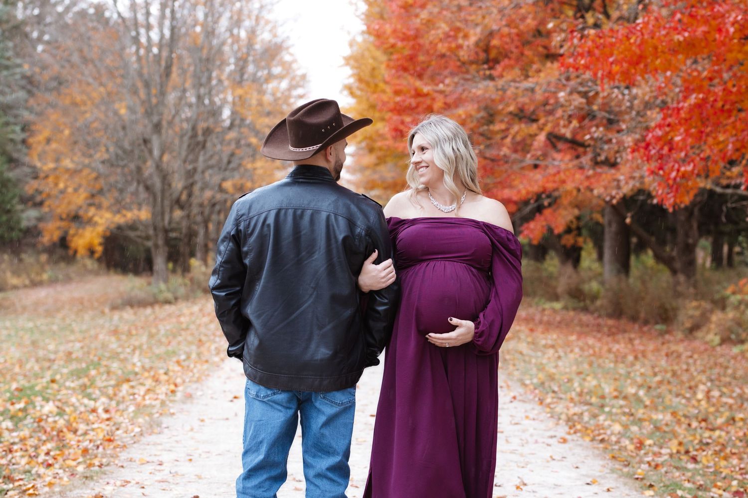 Expectant couple on scenic autumn trail, woman in off-shoulder purple dress cradling baby bump.