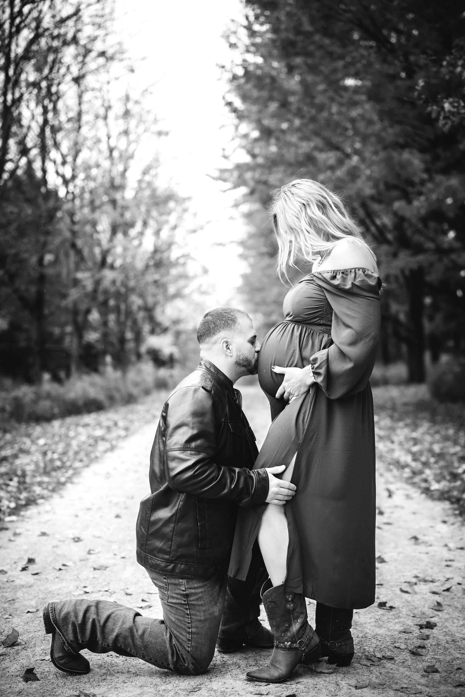 Black and white photo of man kneeling kissing pregnant womans belly outdoors.
