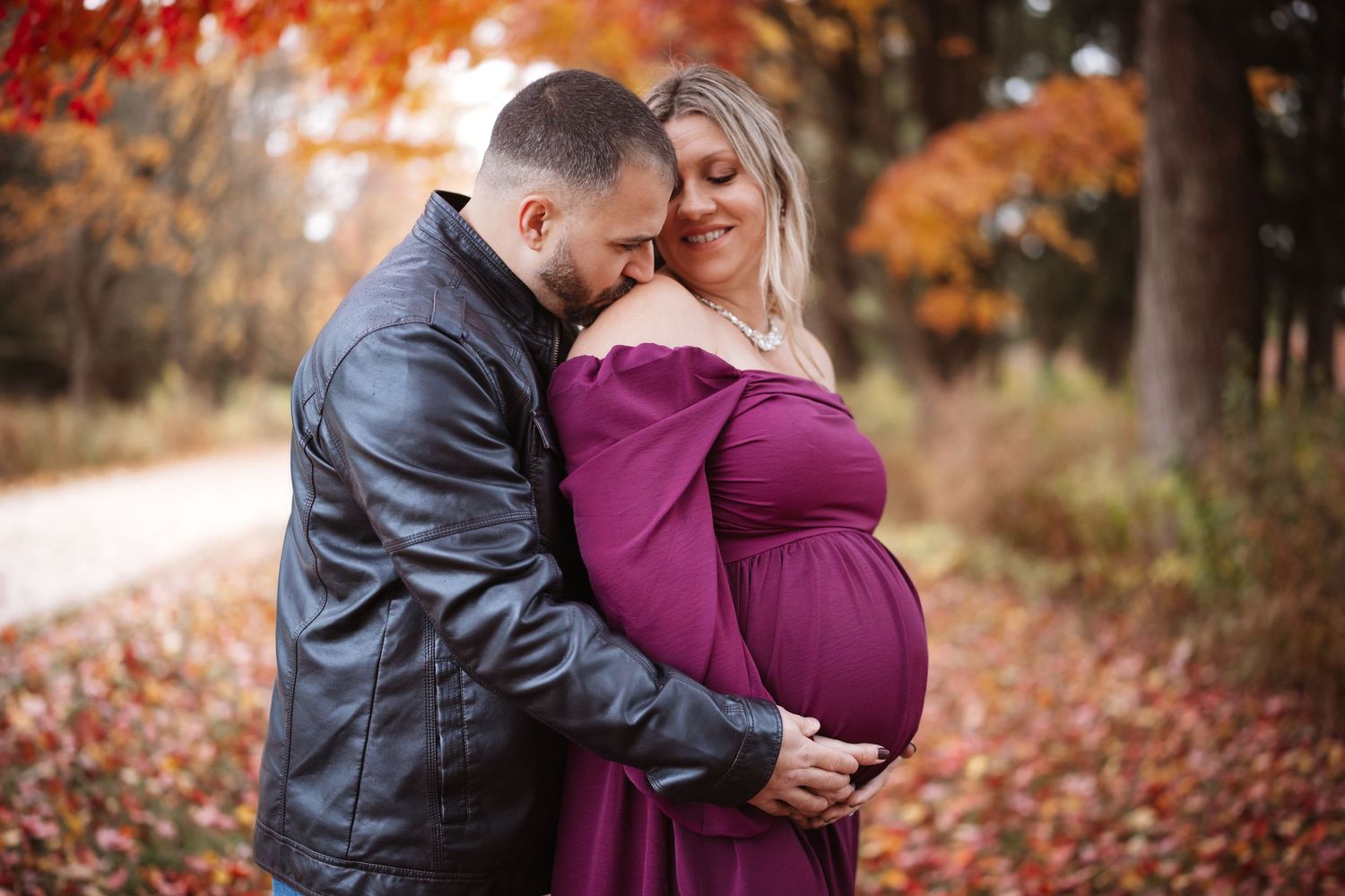 Couple embracing in autumn forest, pregnant woman in purple maternity gown with partner in leather jacket.