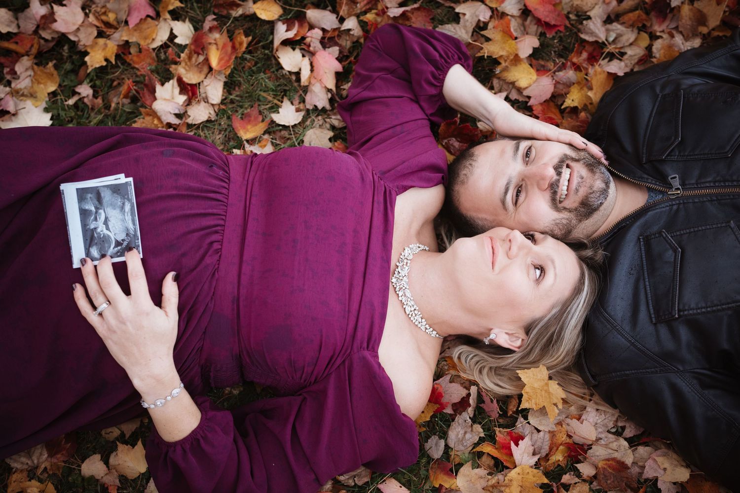Happy couple lying in autumn leaves, pregnant woman in purple dress holding ultrasound photo, both smiling.