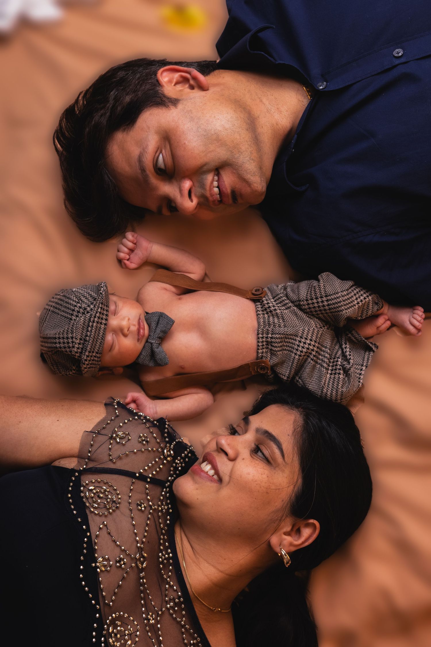 Happy parents lying down smiling at newborn baby in plaid newsboy hat between them.