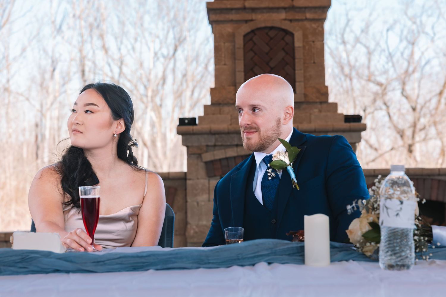 Bride and groom seated at wedding reception table with candles and stone fireplace backdrop in winter setting.