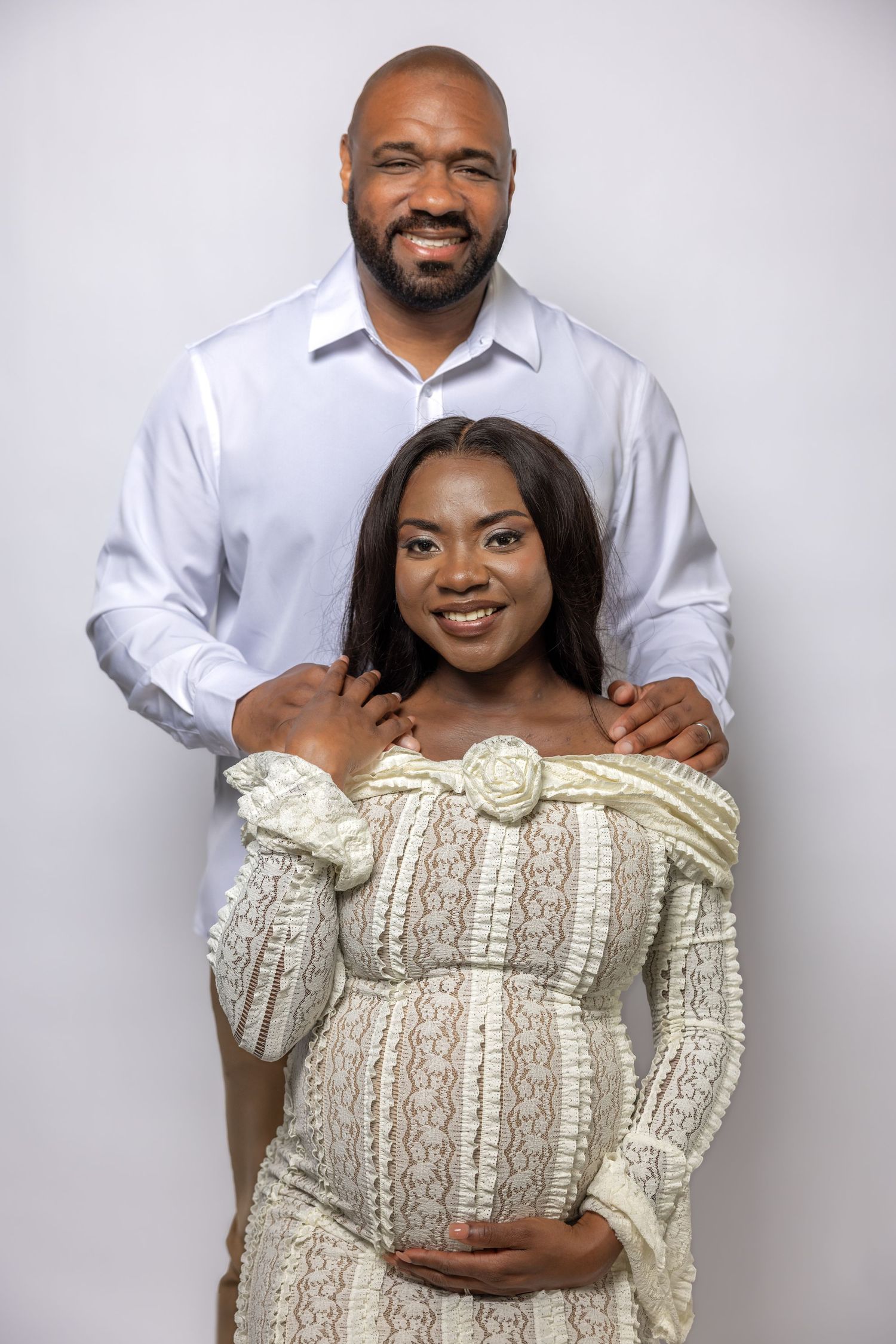 Happy expecting couple posing together, woman in cream lace maternity gown cradling baby bump.