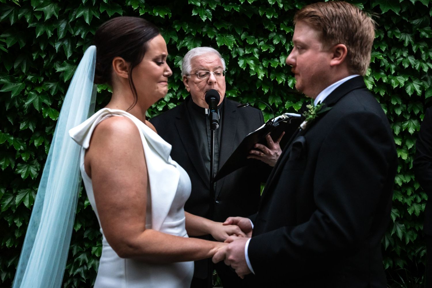 Bride and groom hold hands while exchanging vows with officiant during outdoor wedding ceremony.