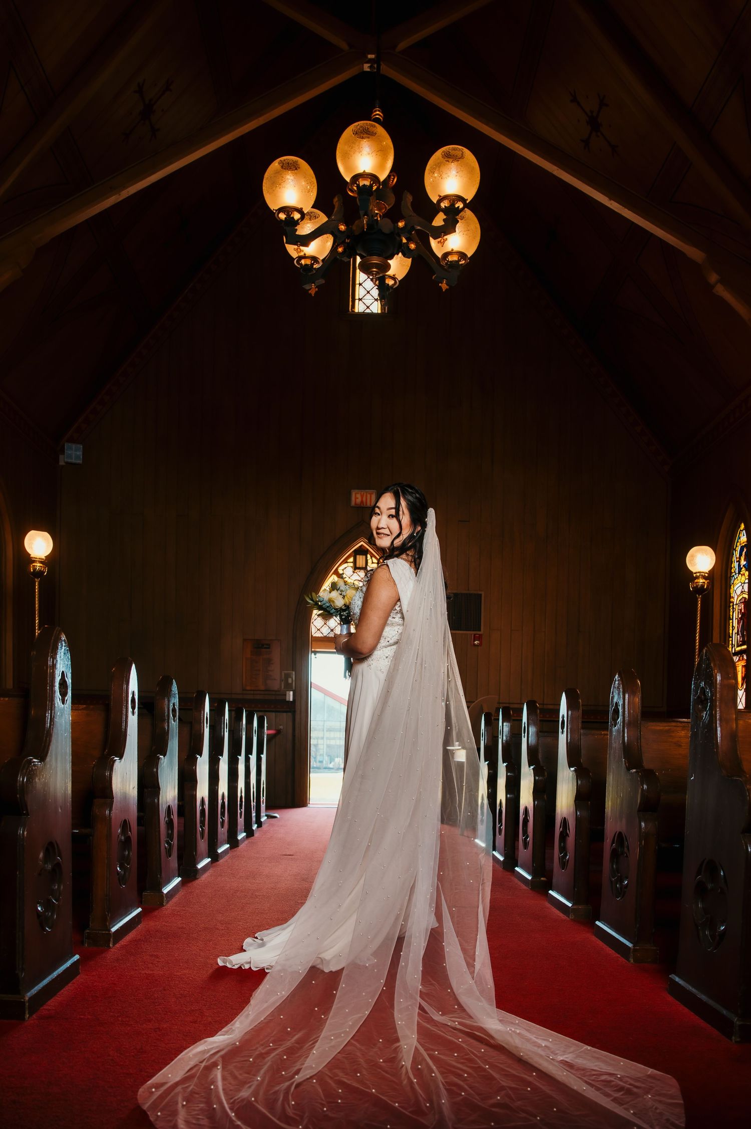 Elegant bride in white gown with long cathedral veil standing in aisle of wooden chapel.