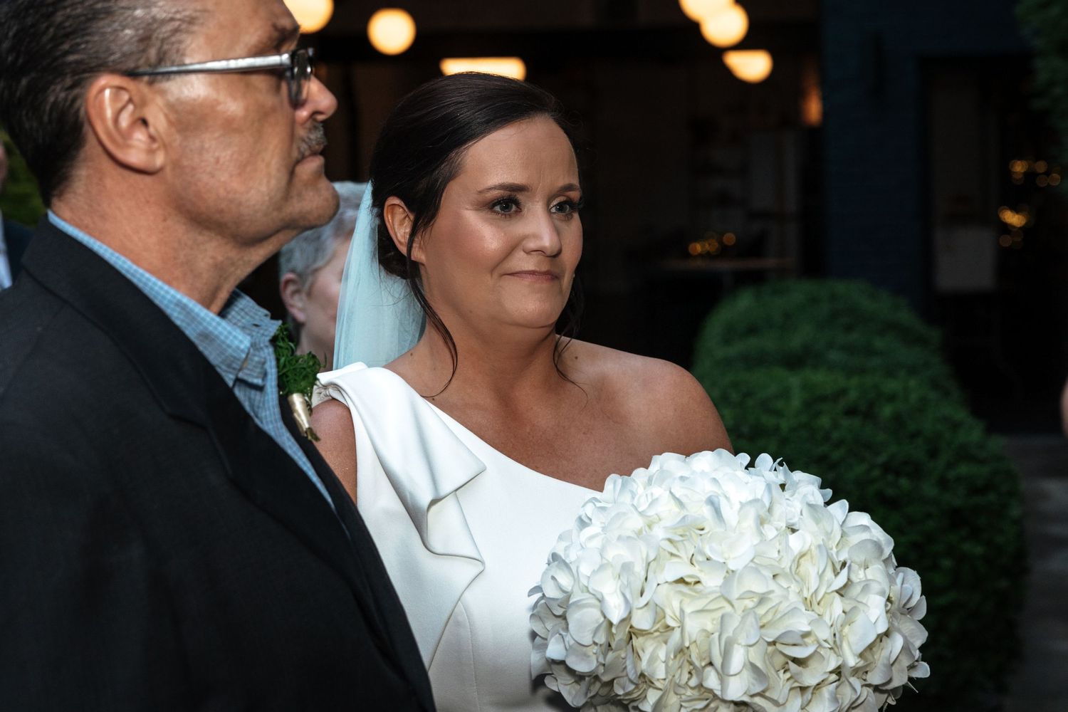 Bride in elegant white wedding dress holding large white hydrangea bouquet with escort at outdoor ceremony.