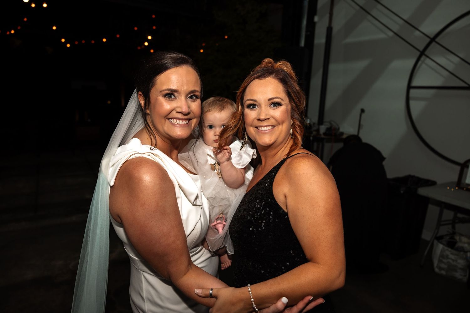 Bride in white gown holds baby in white dress beside woman in black at outdoor evening wedding.