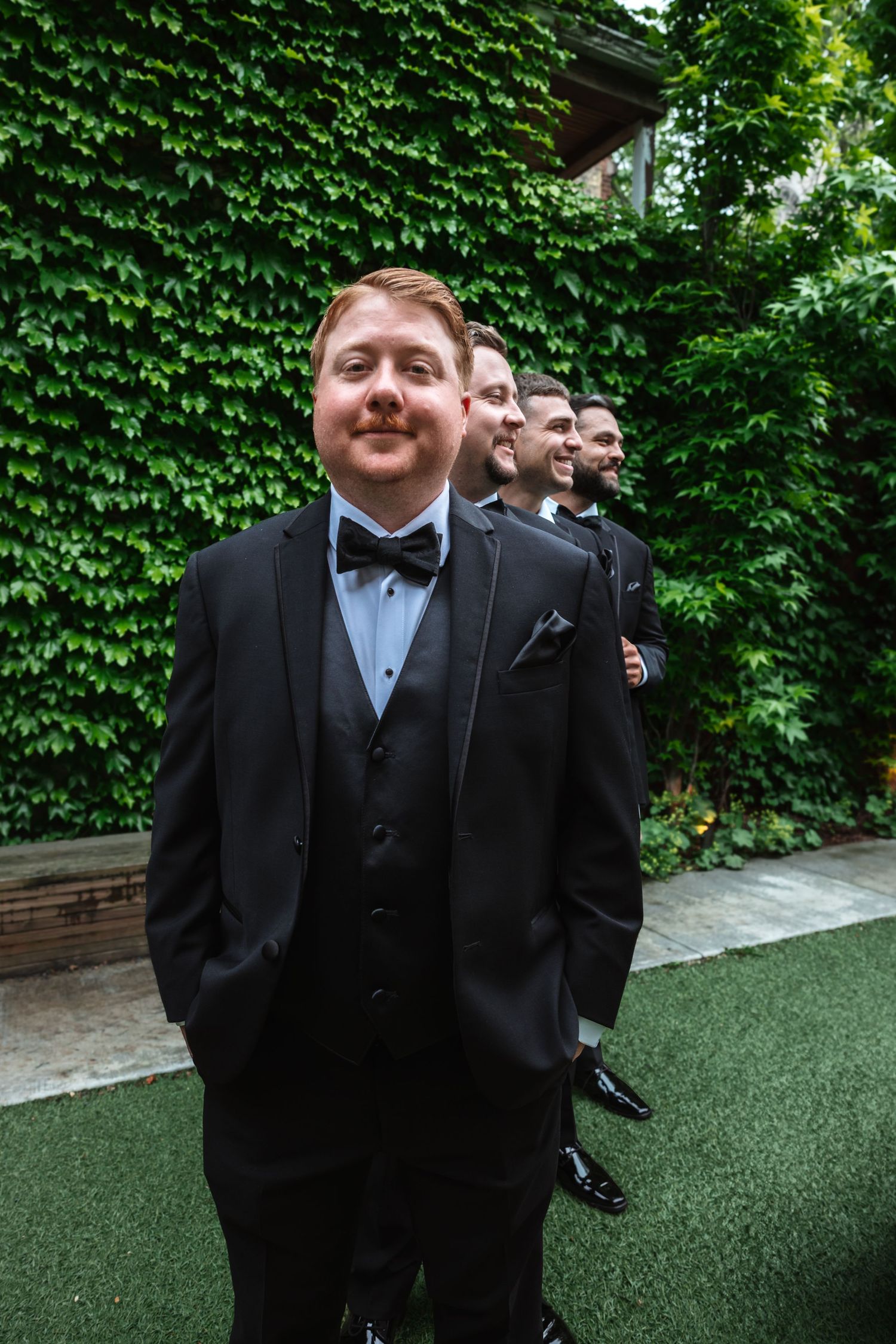 Groom in black tuxedo and bow tie standing confidently with smiling groomsmen lined up behind him outdoors.