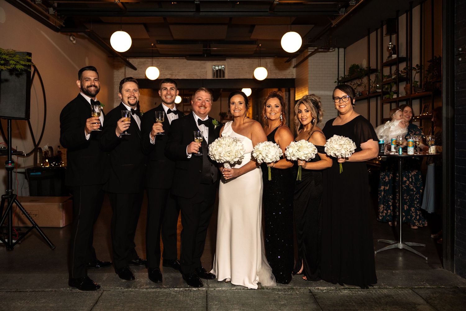Wedding party in black formal attire poses with bouquets and champagne at indoor reception venue.