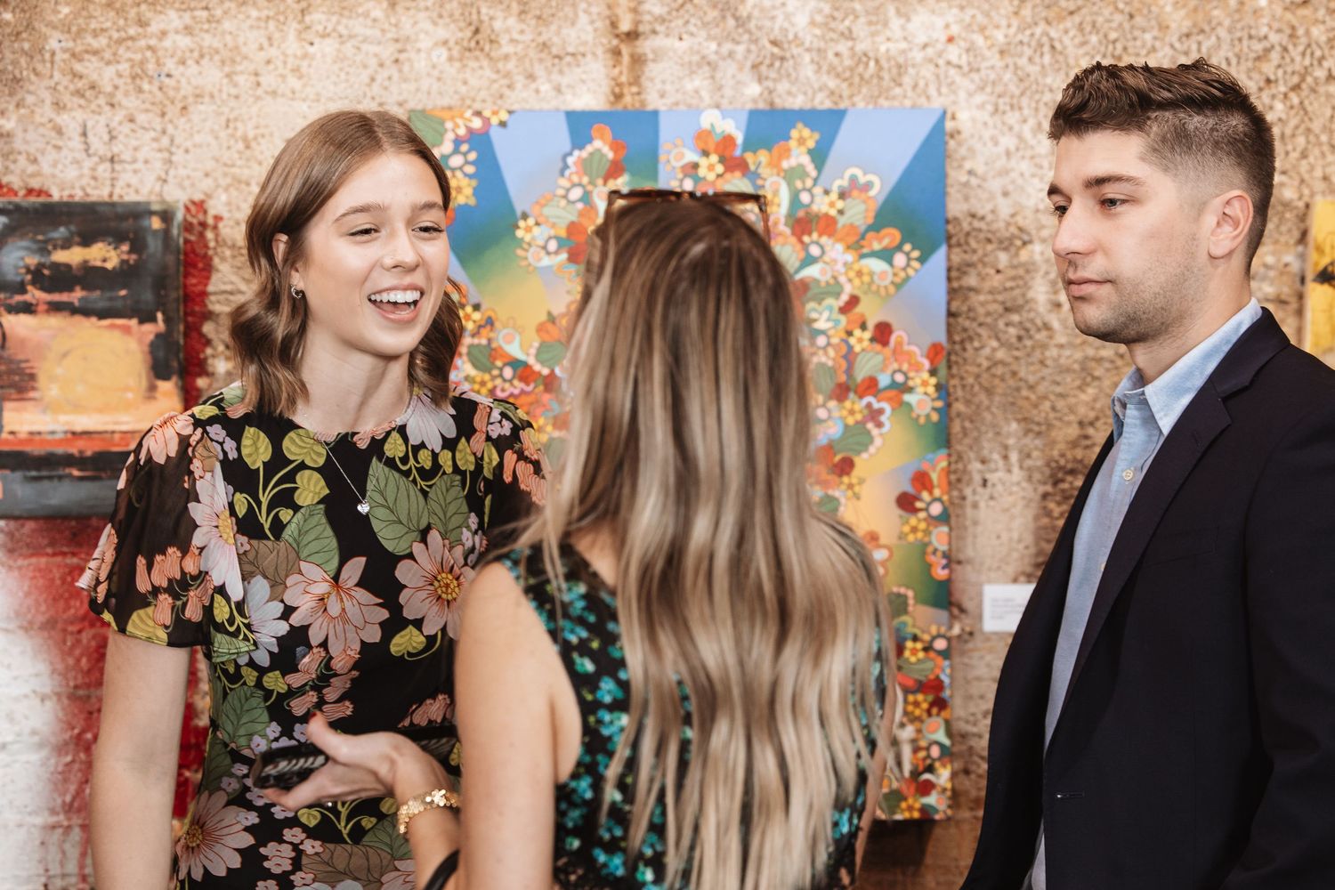 Three young guests admiring colorful floral artwork at an indoor gallery reception.