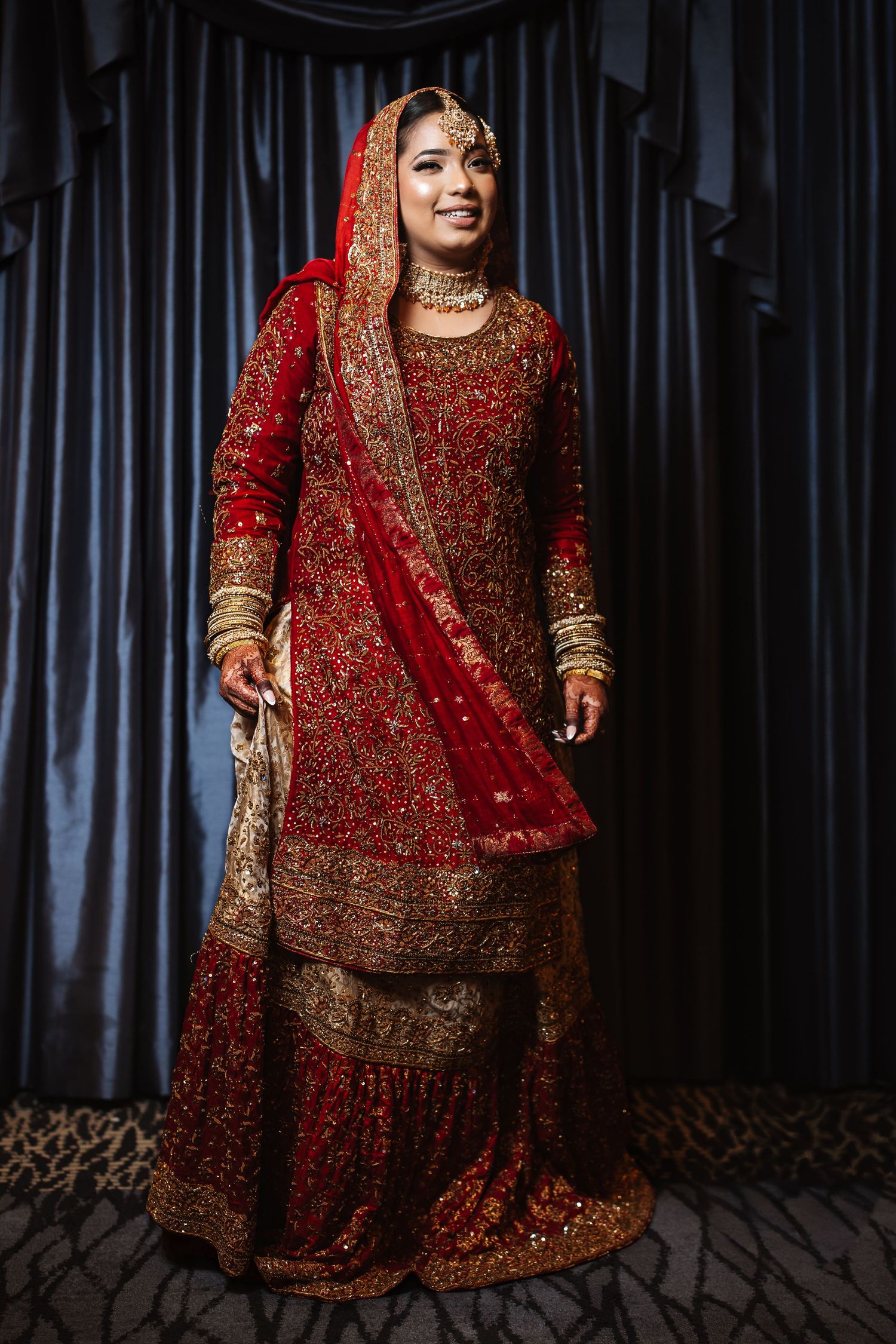 South Asian bride smiling in traditional red and gold embroidered bridal lehenga with dupatta and jewelry.