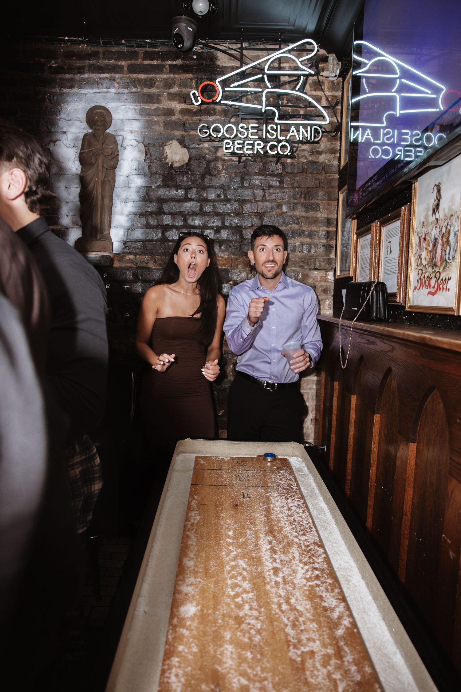 Excited couple playing shuffleboard at Goose Island Beer Co bar during wedding reception.