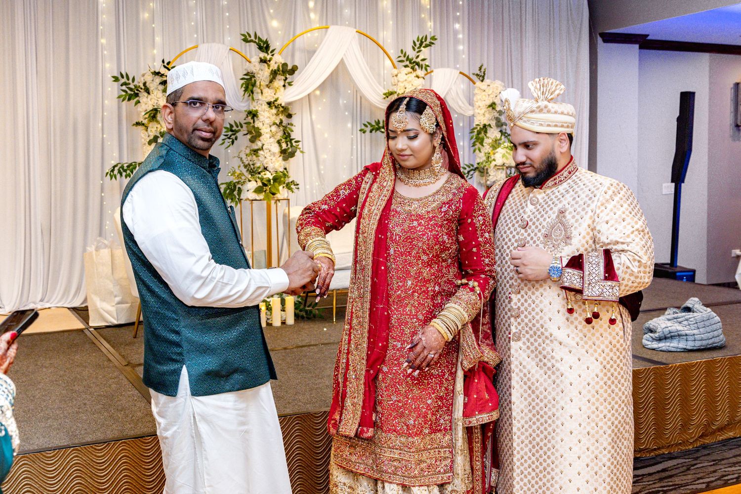 A man in teal vest shakes hands with a South Asian groom while bride stands nearby at wedding.