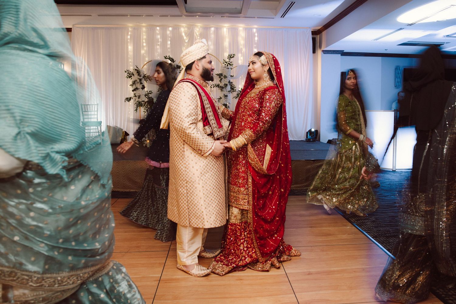 South Asian bride and groom in traditional red and gold wedding attire hold hands during ceremony.