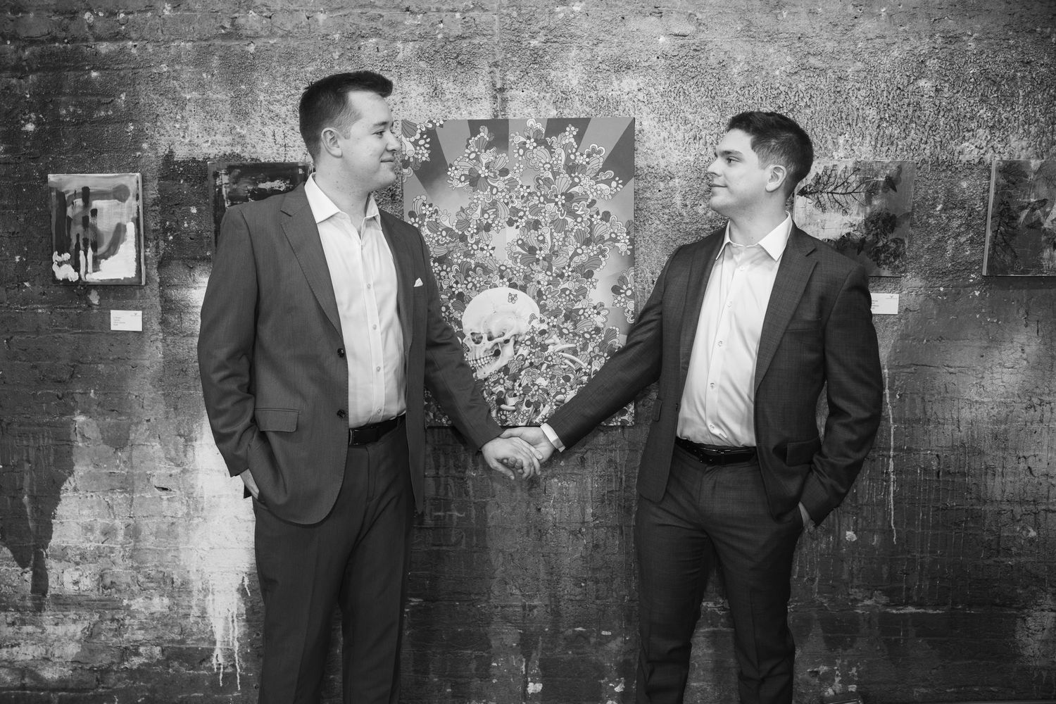 Black and white photo of two suited men holding hands in front of artwork on a brick wall.