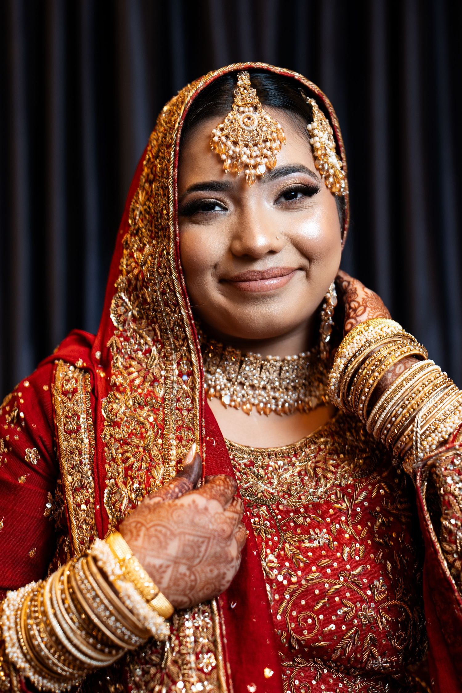 Smiling Indian bride in red lehenga with gold jewelry, bangles, and mehndi on her hands.