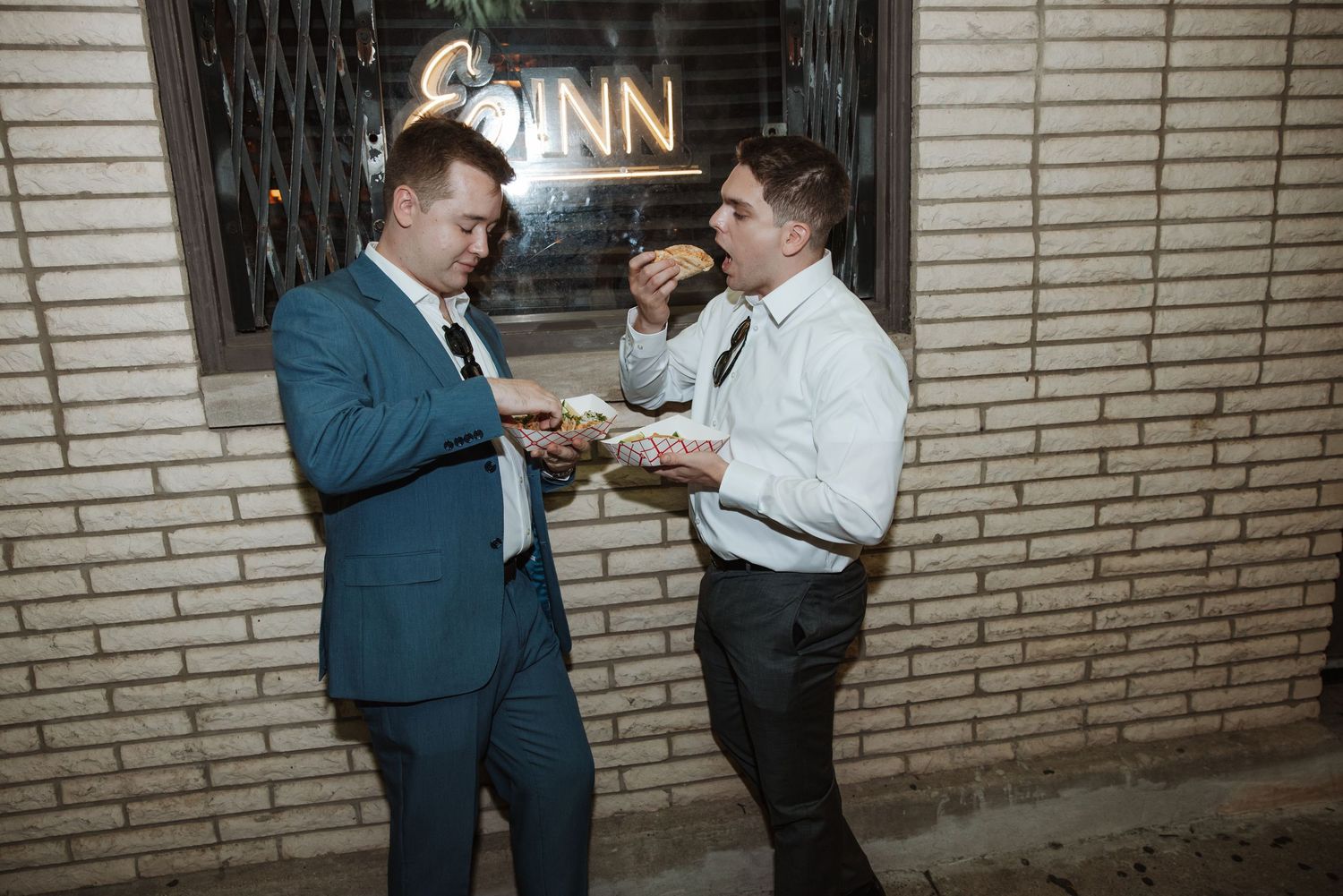 Two men in suits share food outside a brick building at night.