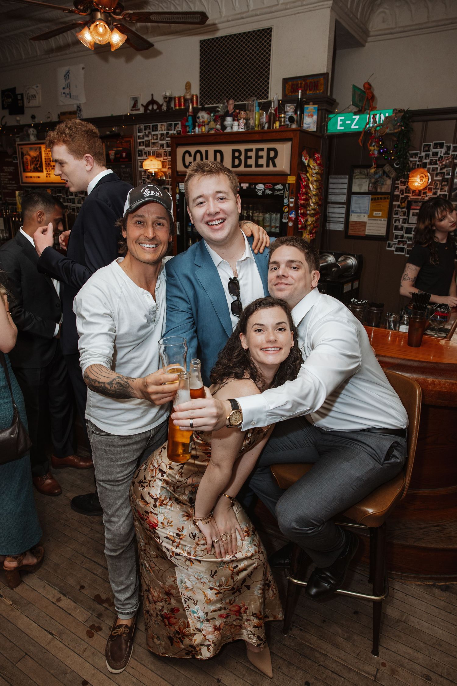 Four friends smile and clink beer glasses at a rustic bar with a Cold Beer sign.
