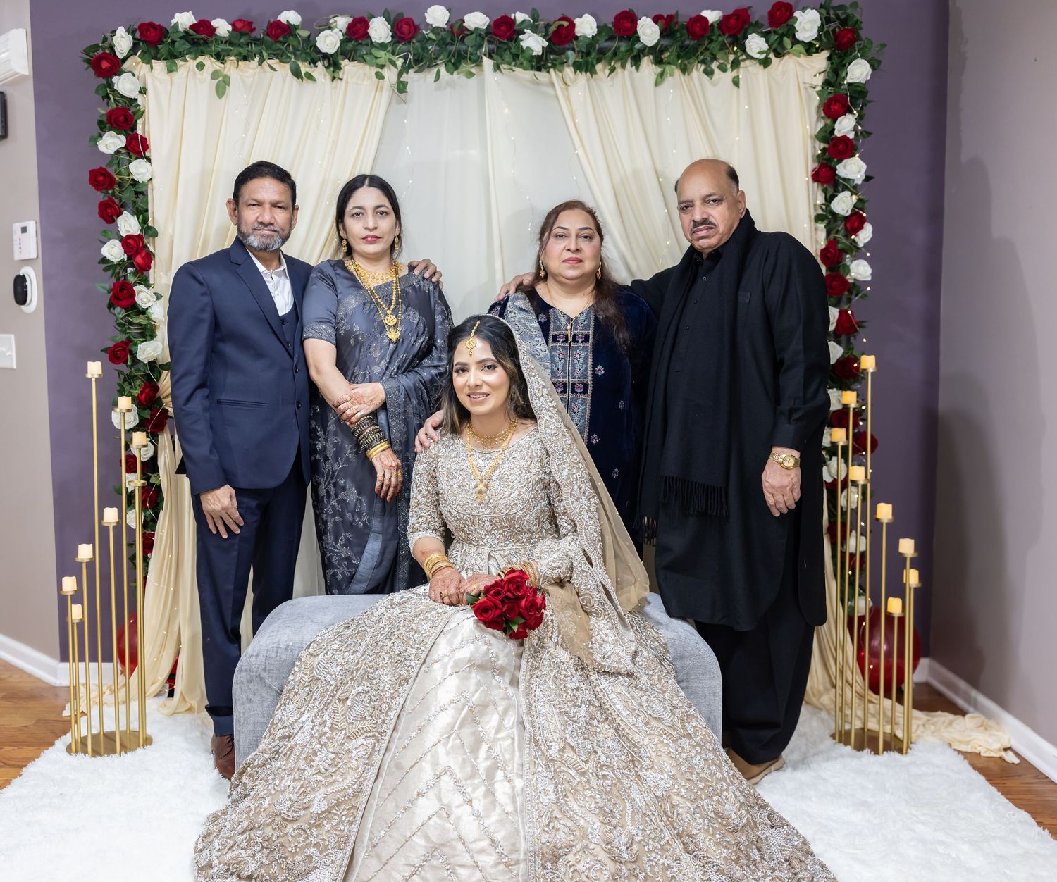 Bride in embroidered gold lehenga poses with four family members before floral wedding arch backdrop.