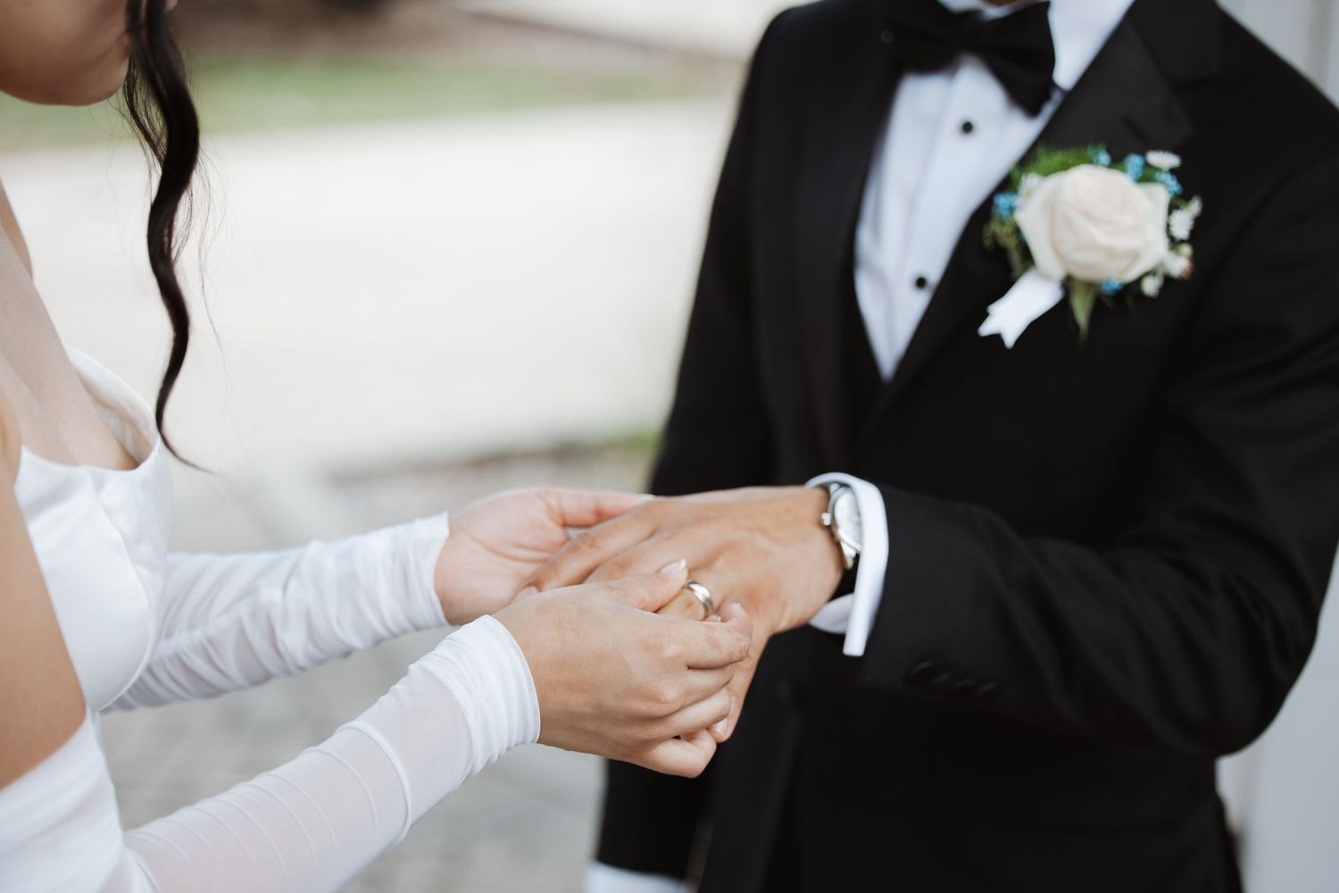 Bride and groom exchanging rings outdoors, groom in black tuxedo with white boutonniere.