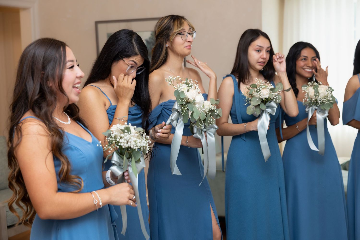 Emotional bridesmaids in blue dresses cry and smile while holding white bouquets during bridal reveal.