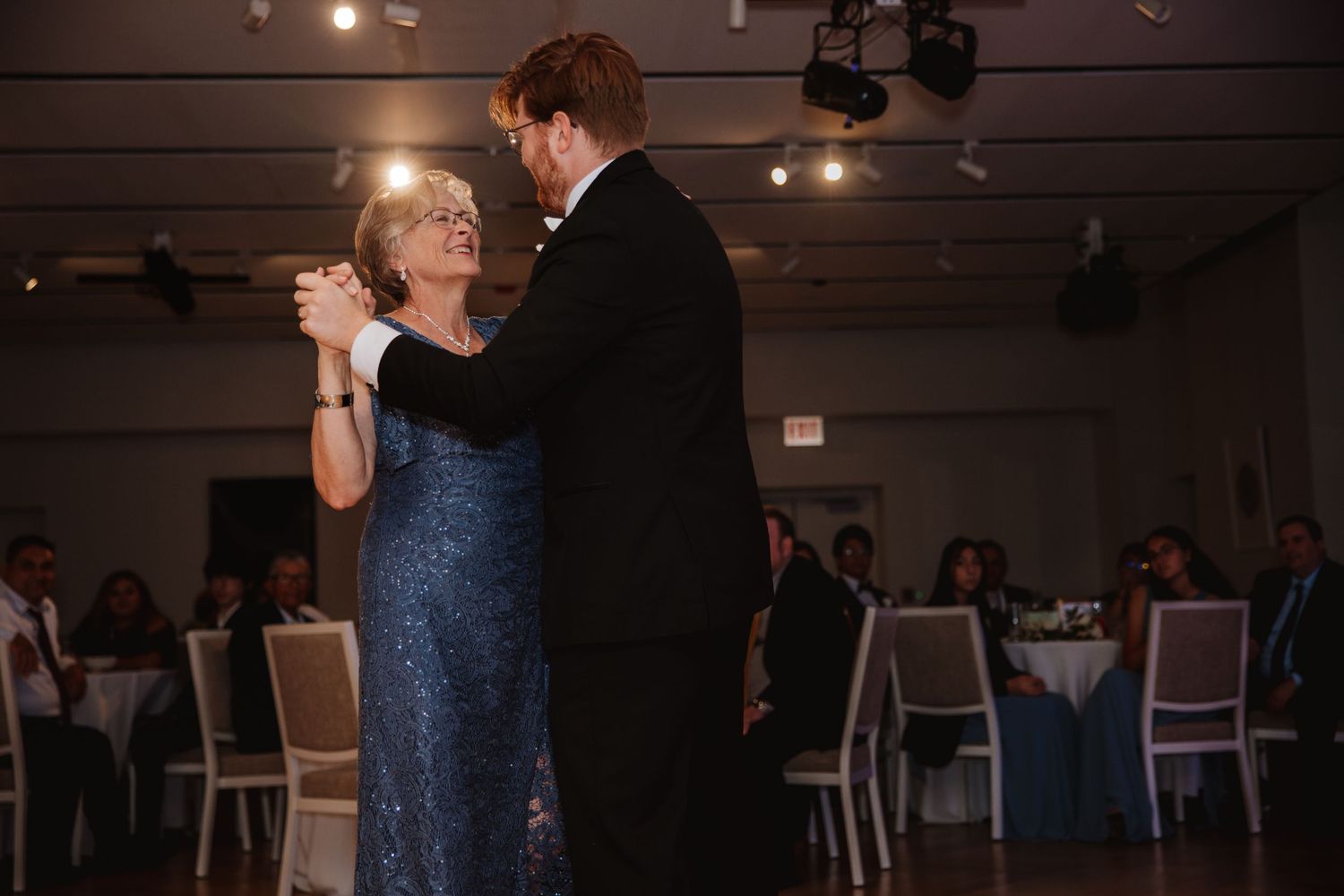 Groom in black tuxedo shares emotional mother-son dance at elegant wedding reception.