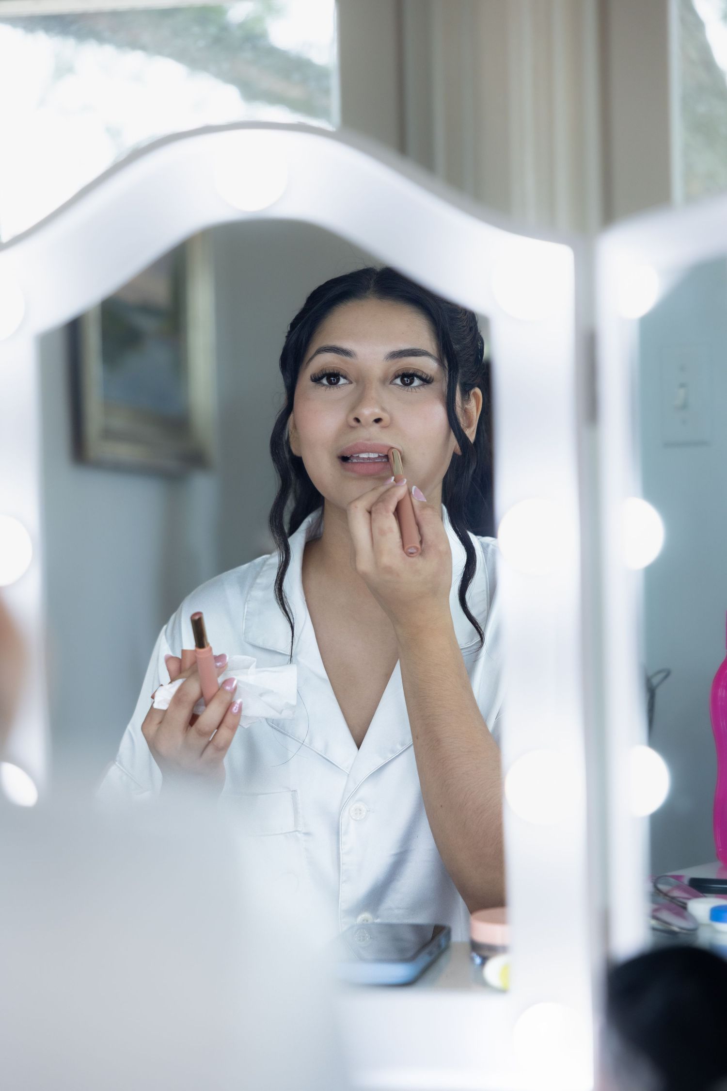 A young Asian woman applies lipstick while gazing into a lighted Hollywood vanity mirror during bridal prep.