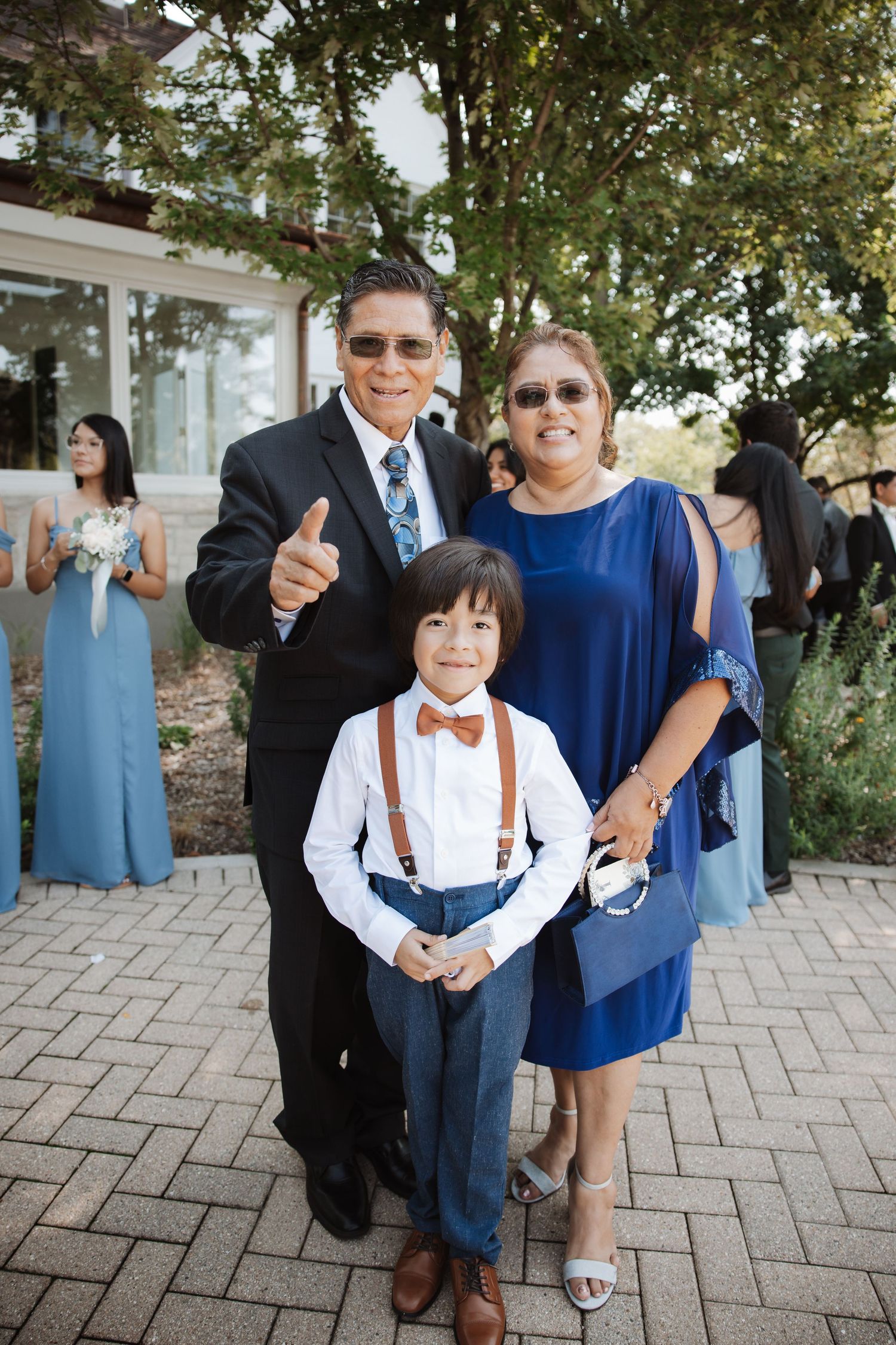 Smiling grandfather gives thumbs up beside grandmother and ring bearer in suspenders at outdoor wedding celebration.