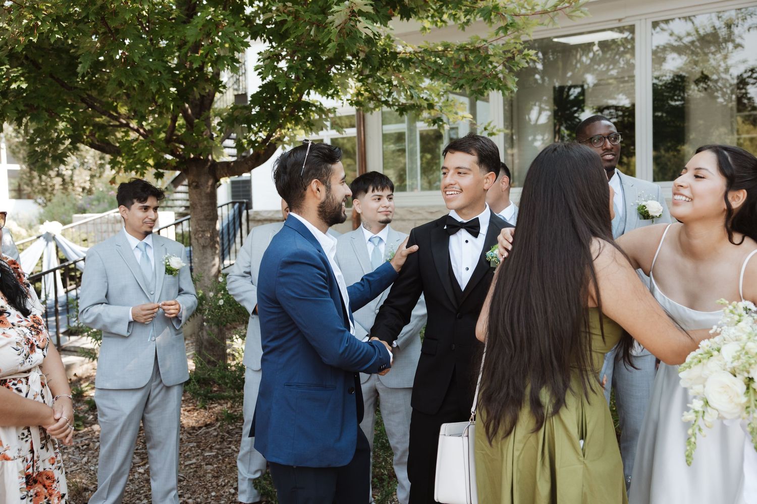 Groom in tuxedo shakes hands with guest while wedding party mingles outdoors after ceremony.