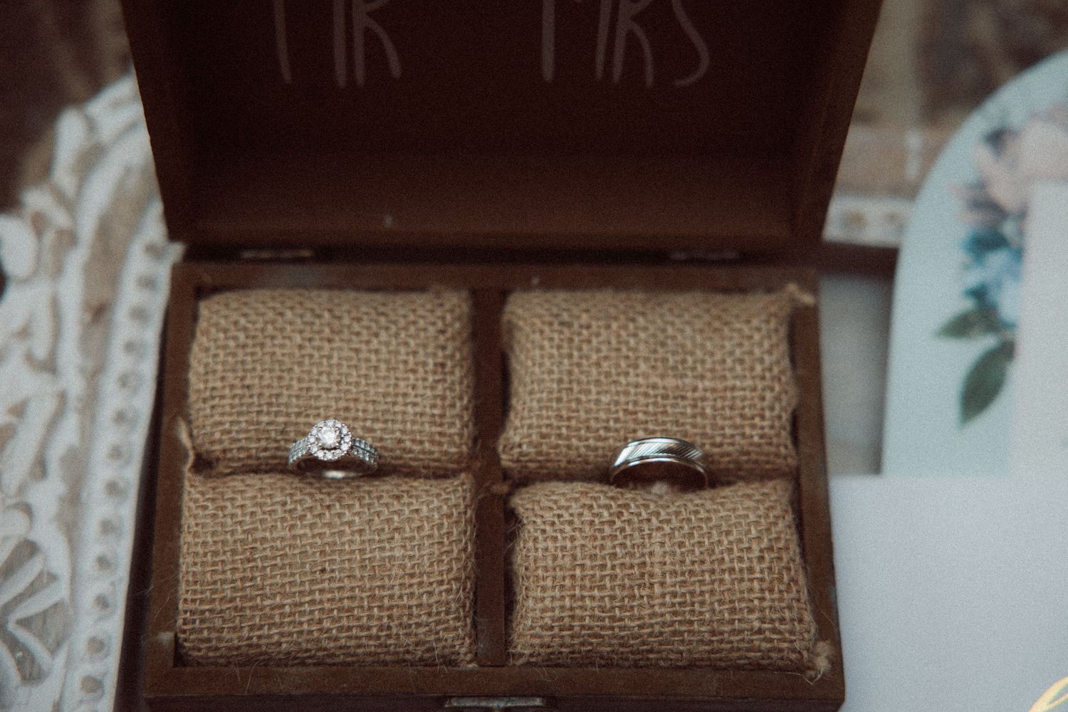 Close-up of wedding rings displayed on burlap cushions inside a rustic Mr and Mrs wooden box.