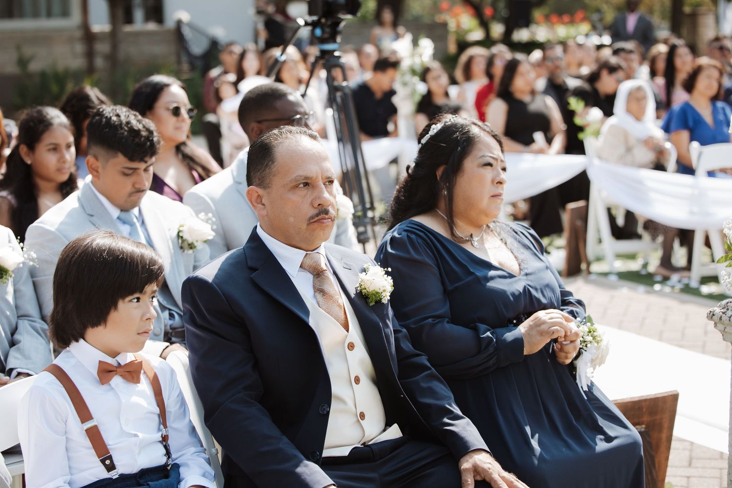 Wedding guests seated outdoors watching ceremony, man in suit and woman in blue dress in foreground.