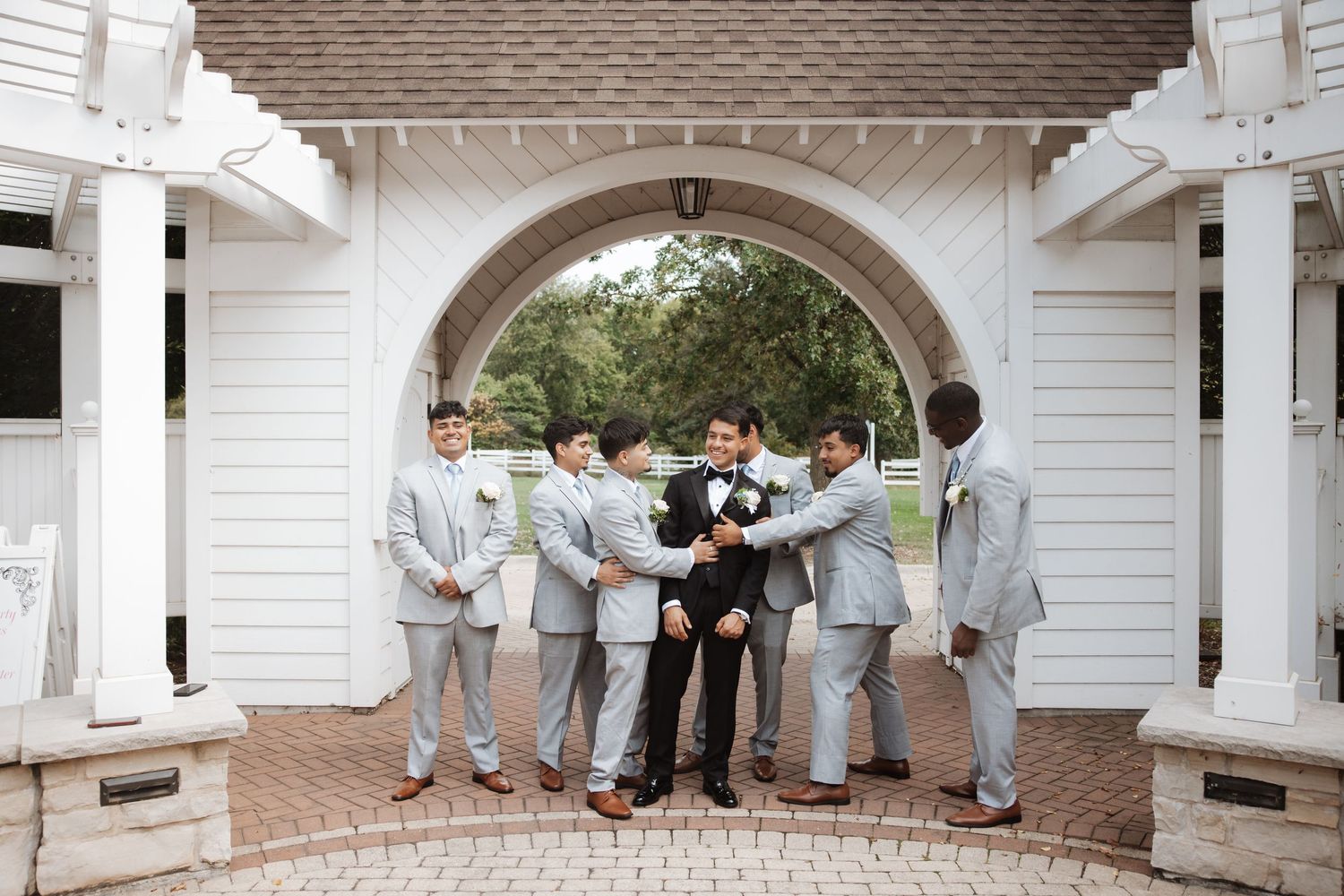 Groomsmen in gray suits gather around groom in black tuxedo under white arch.