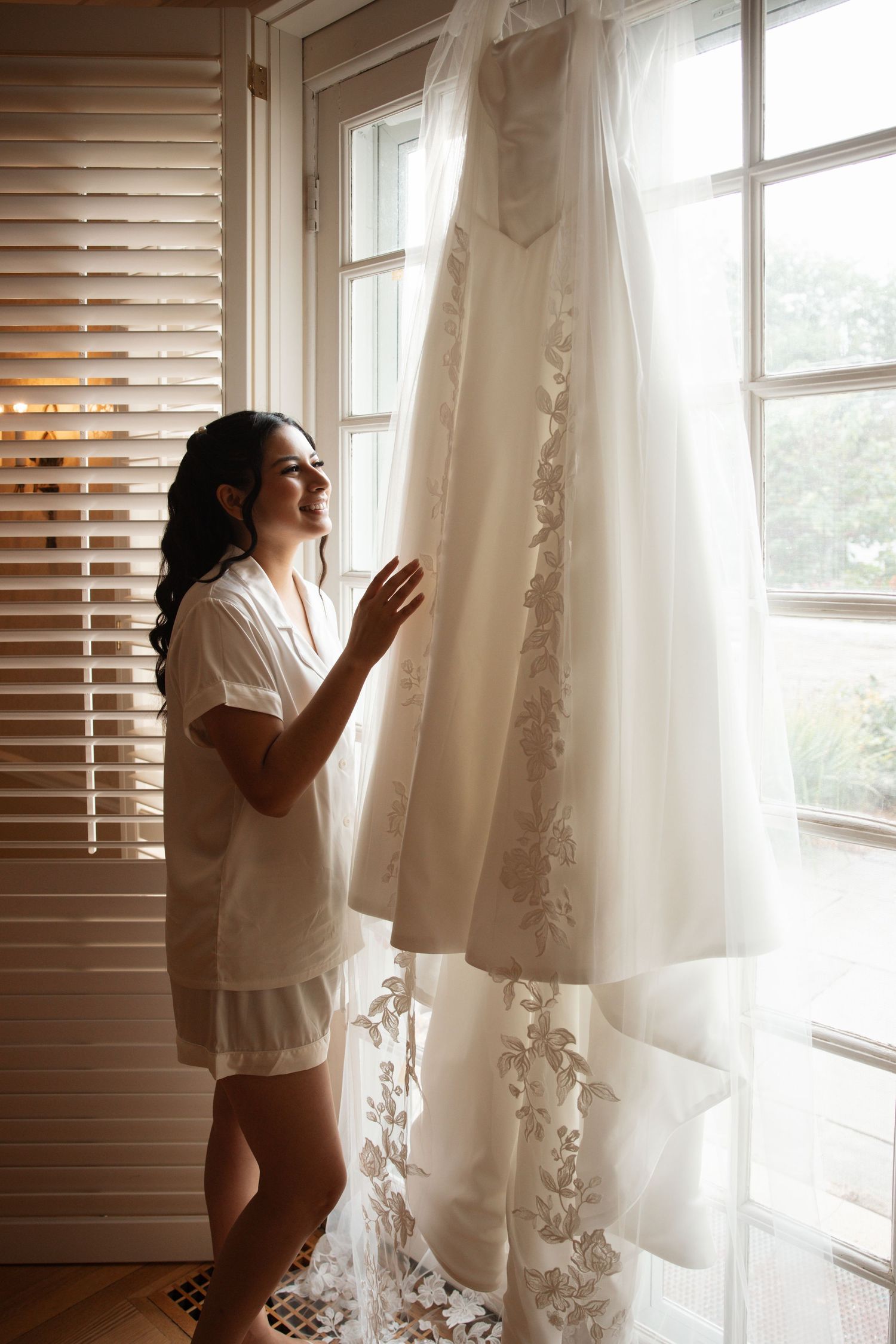 Smiling brunette bride in white robe admiring her lace wedding gown hanging by a bright sunlit window.